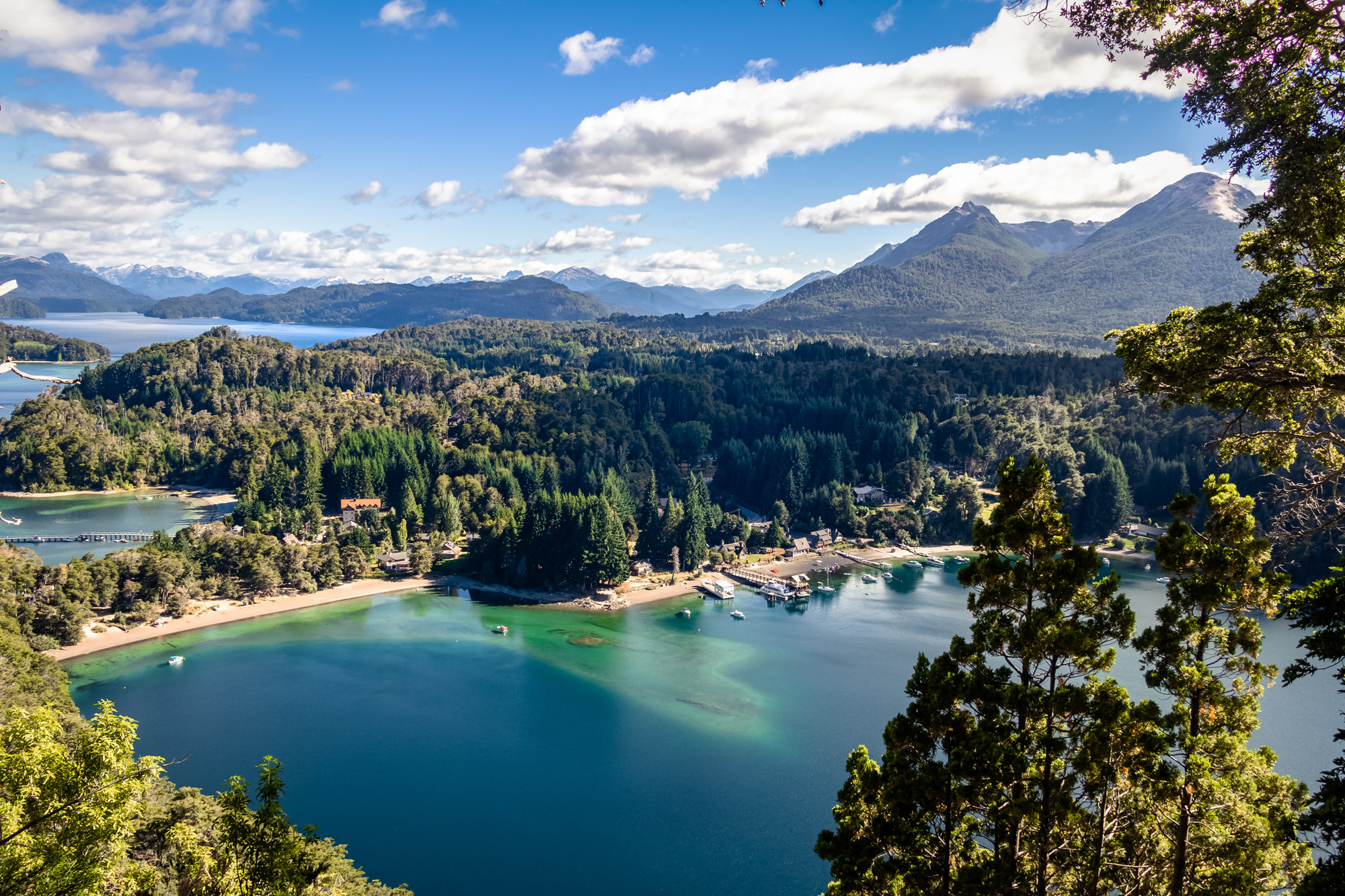 Sunlit blue lake curves through forested hills with rugged mountains in the distance and trees framing the scene.