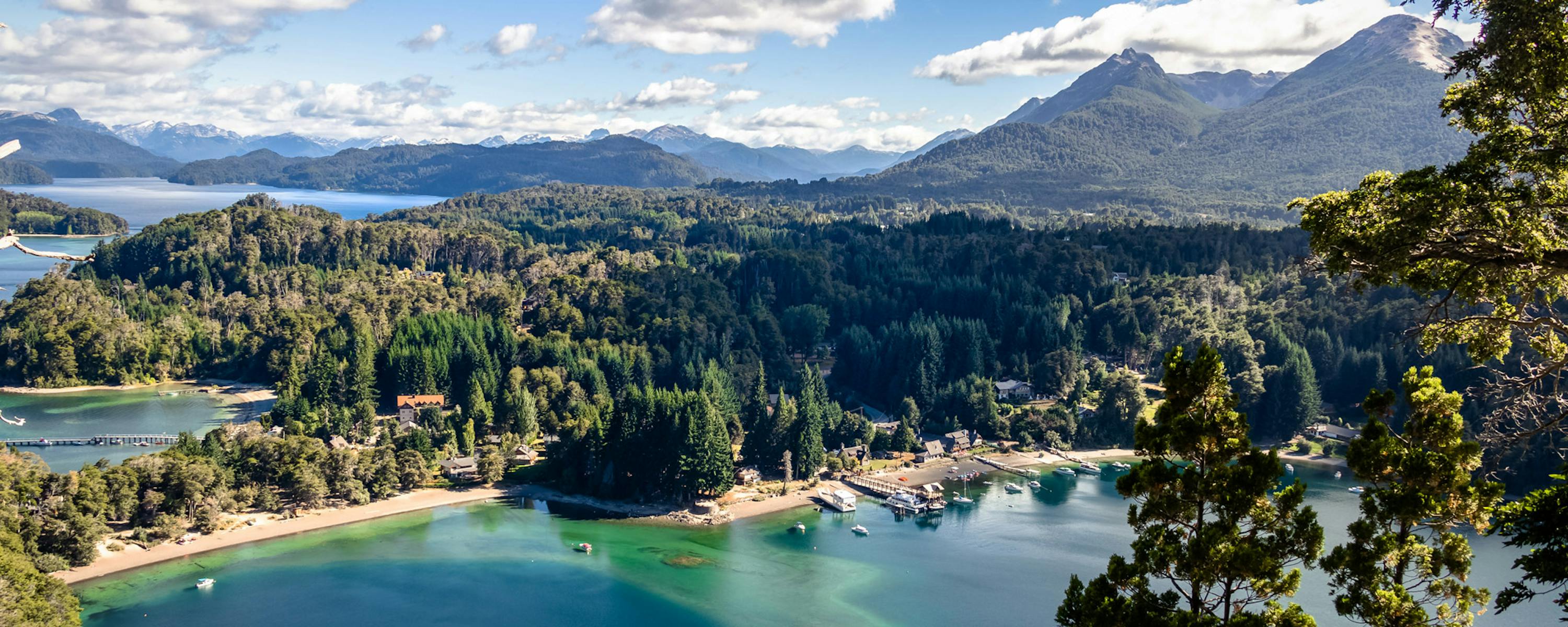 Sunlit blue lake curves through forested hills with rugged mountains in the distance and trees framing the scene.