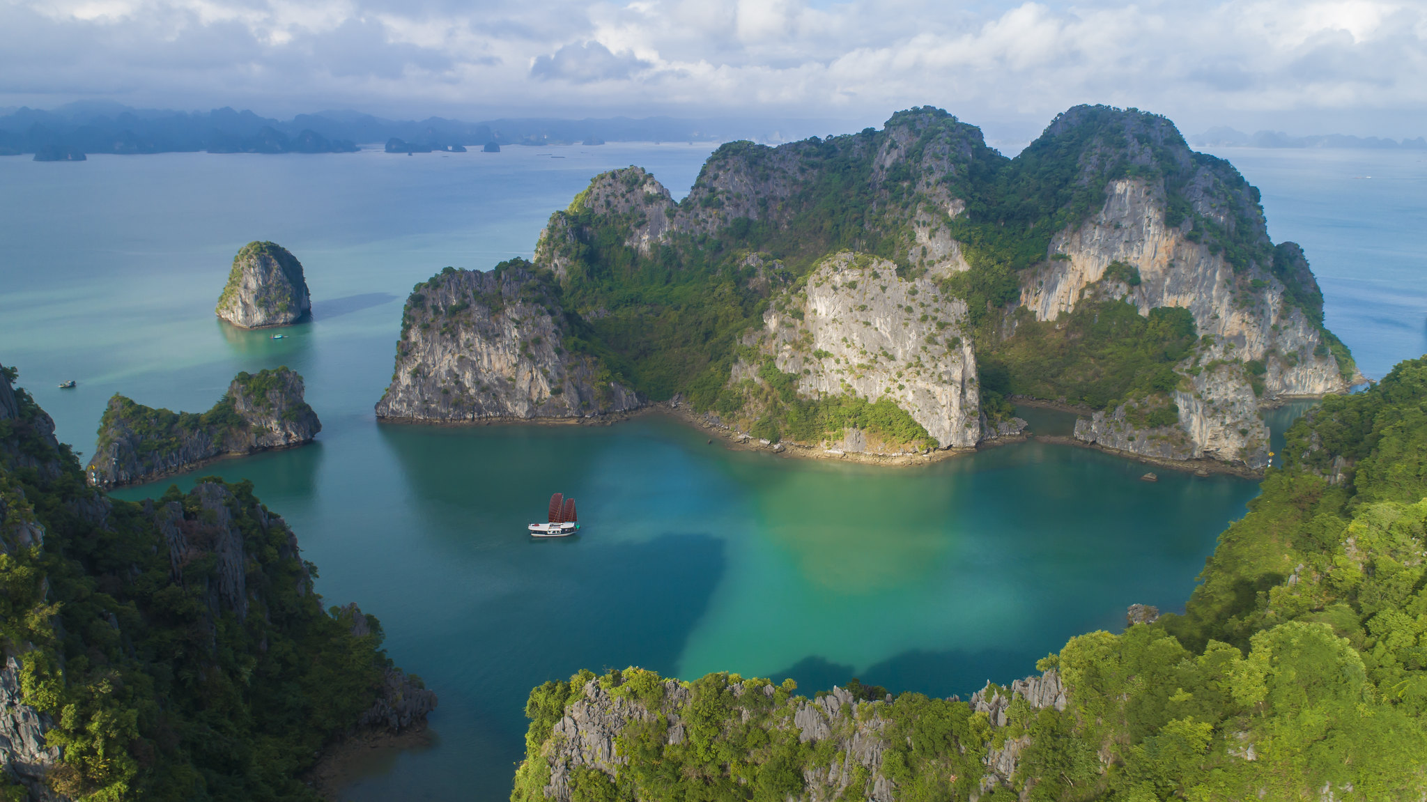 Turquoise water surrounds towering limestone islands, with small boats scattered across the bay under clouds.