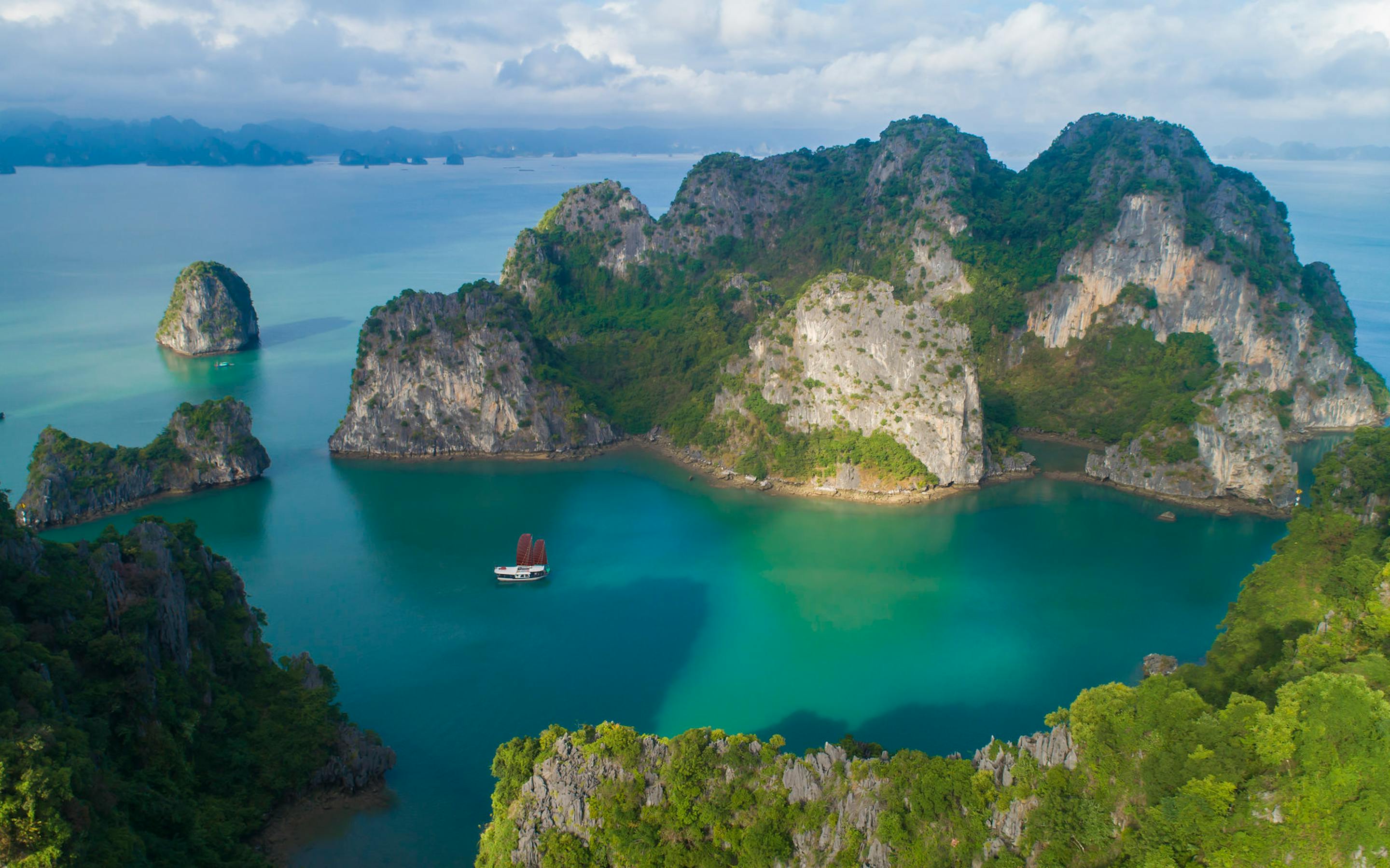 Turquoise water surrounds towering limestone islands, with small boats scattered across the bay under clouds.