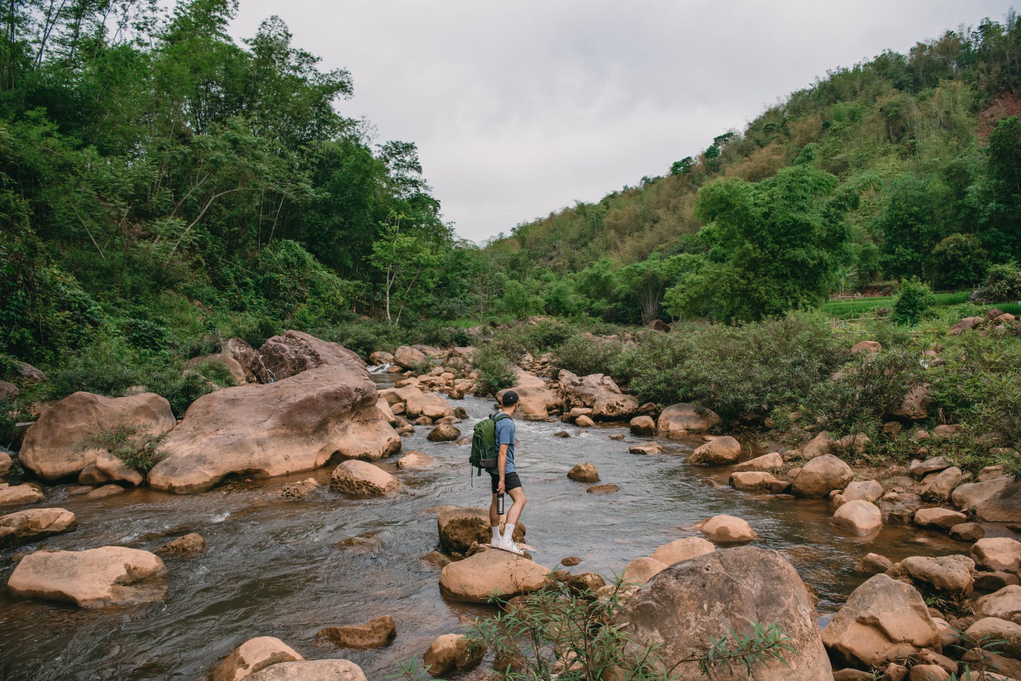 A person steps across a shallow rocky stream, with green hills rising on both sides beneath an overcast sky.