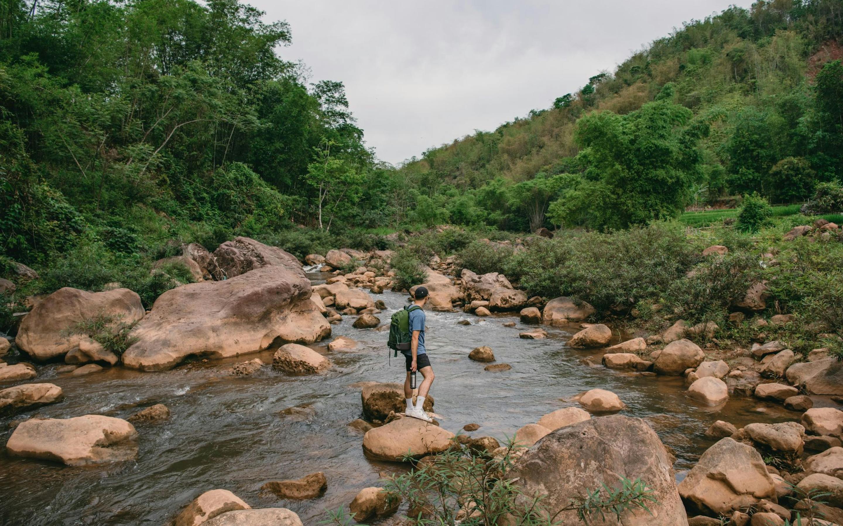 A person steps across a shallow rocky stream, with green hills rising on both sides beneath an overcast sky.