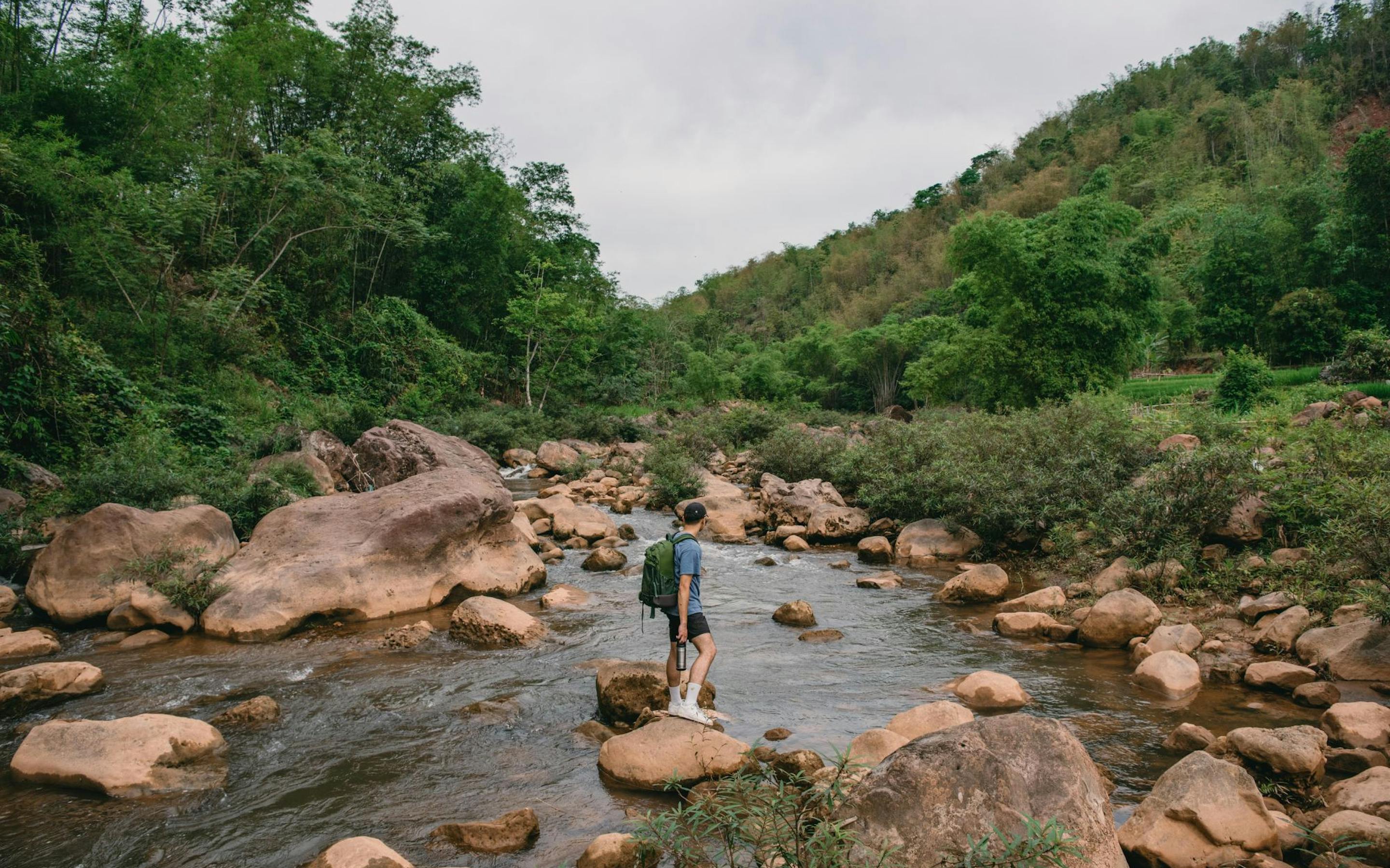 A person steps across a shallow rocky stream, with green hills rising on both sides beneath an overcast sky.