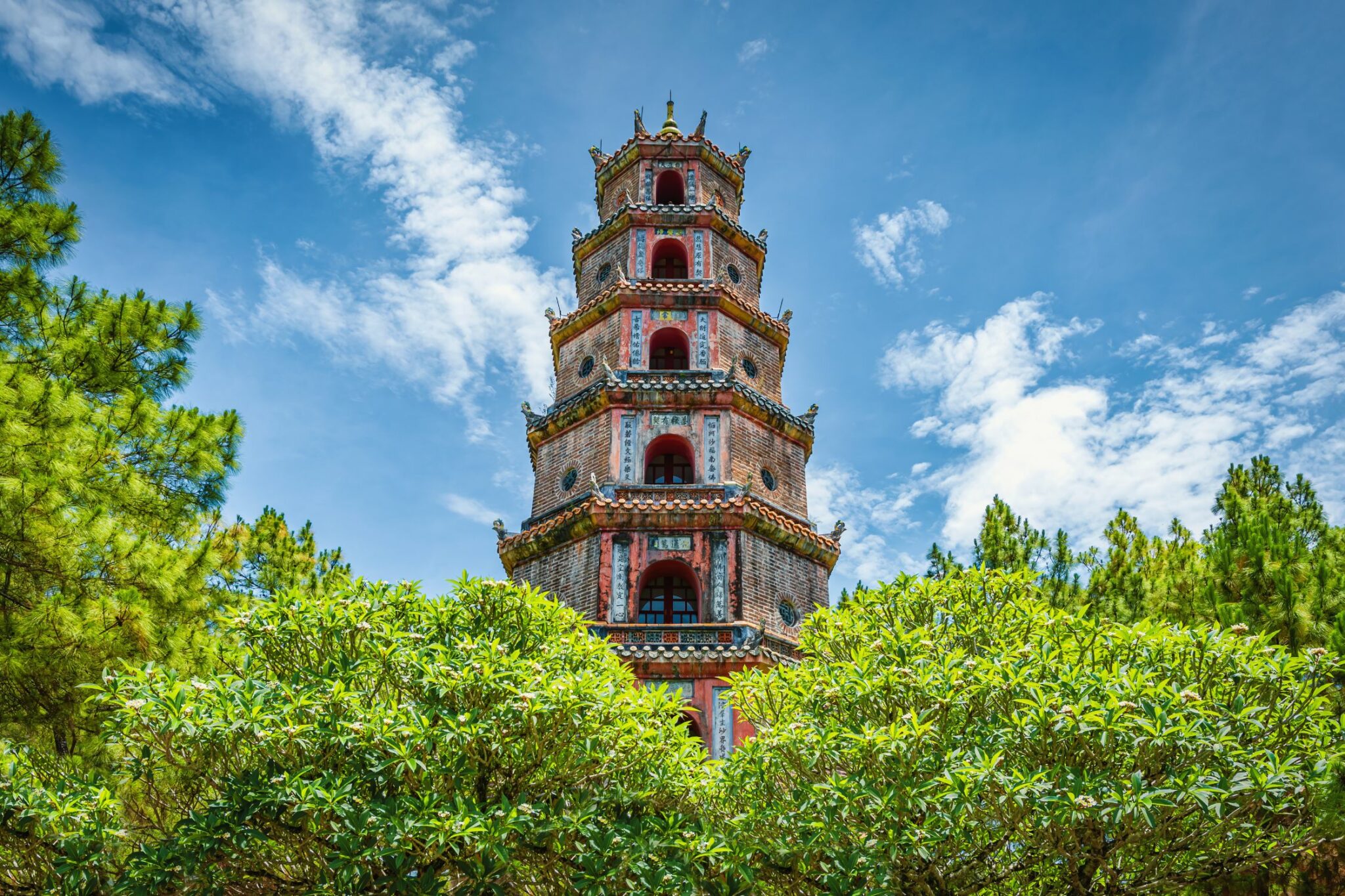 A tall multi-tier pagoda rises above lush green trees, set against a bright blue sky with scattered clouds.