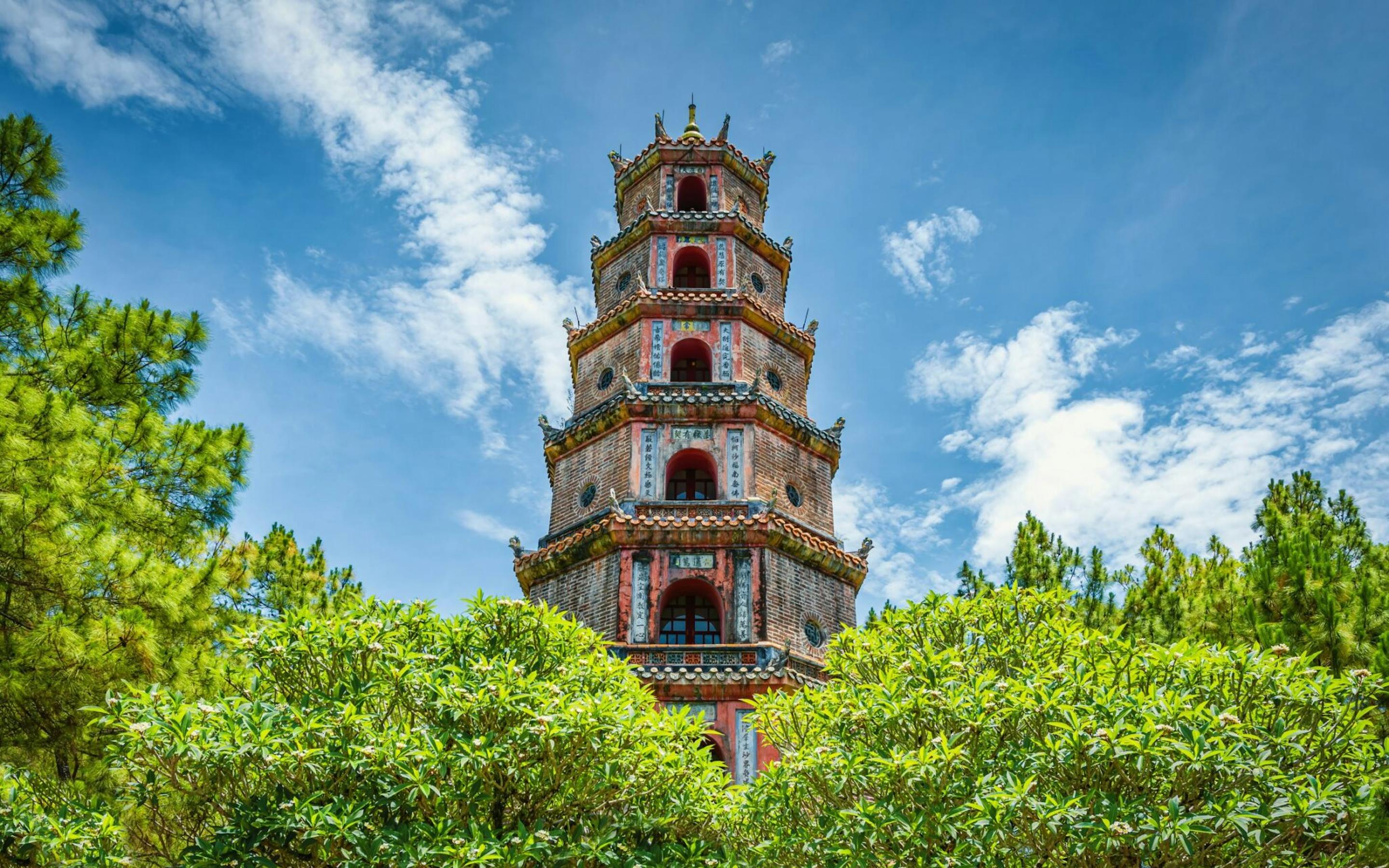 A tall multi-tier pagoda rises above lush green trees, set against a bright blue sky with scattered clouds.