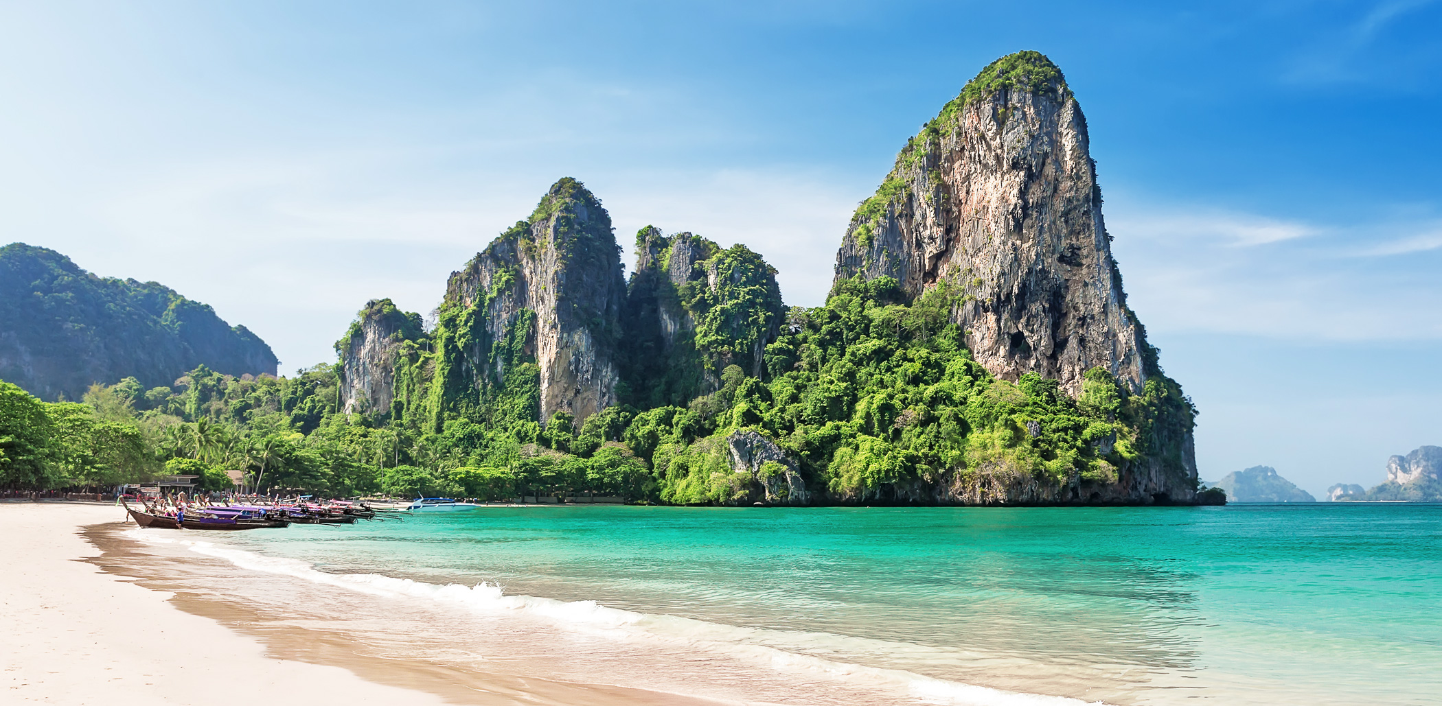Limestone karst cliffs rise above Railay Beach, where pale sand meets clear turquoise water in bright sun.