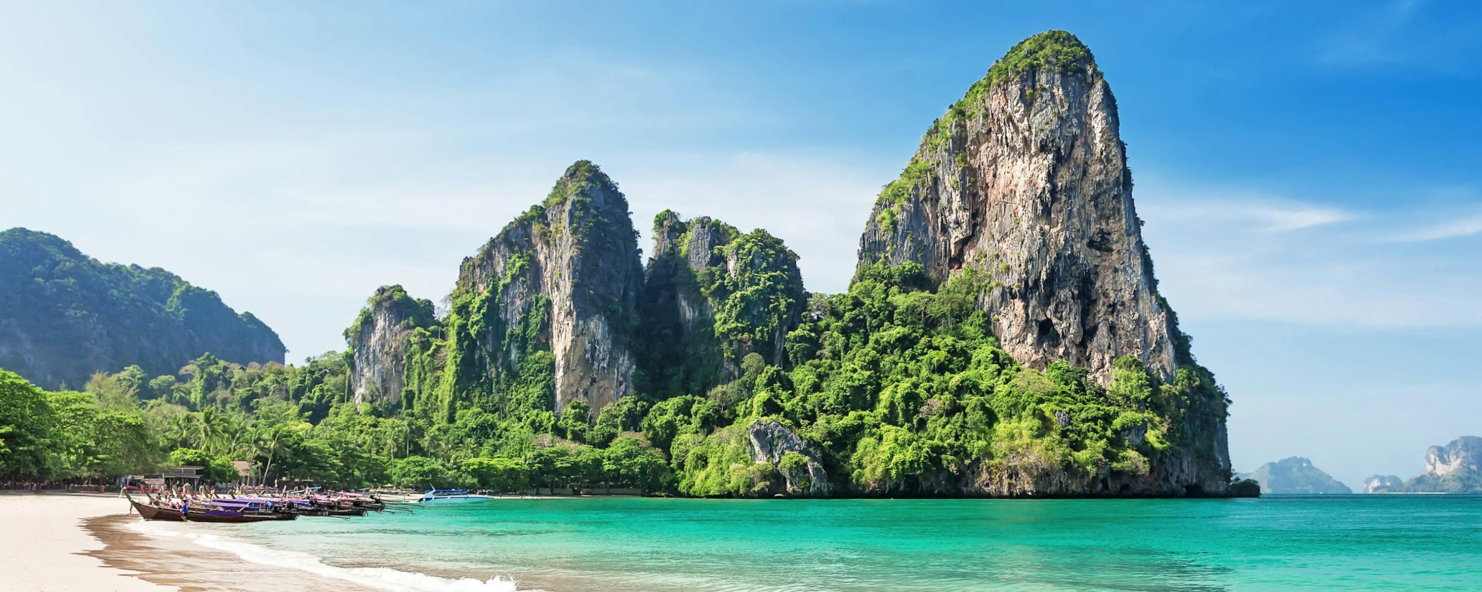 Limestone karst cliffs rise above Railay Beach, where pale sand meets clear turquoise water in bright sun.