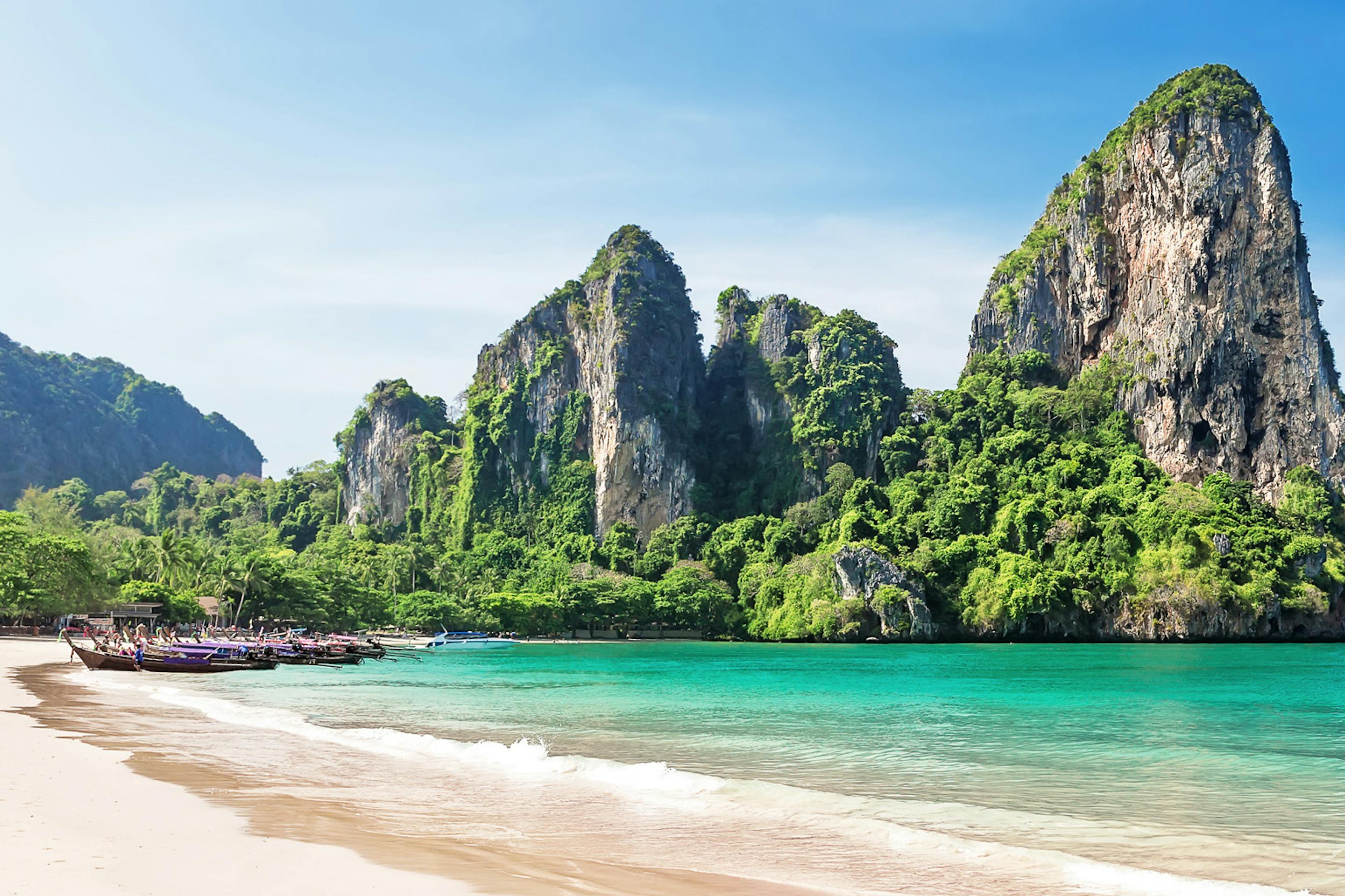 Limestone karst cliffs rise above Railay Beach, where pale sand meets clear turquoise water in bright sun.