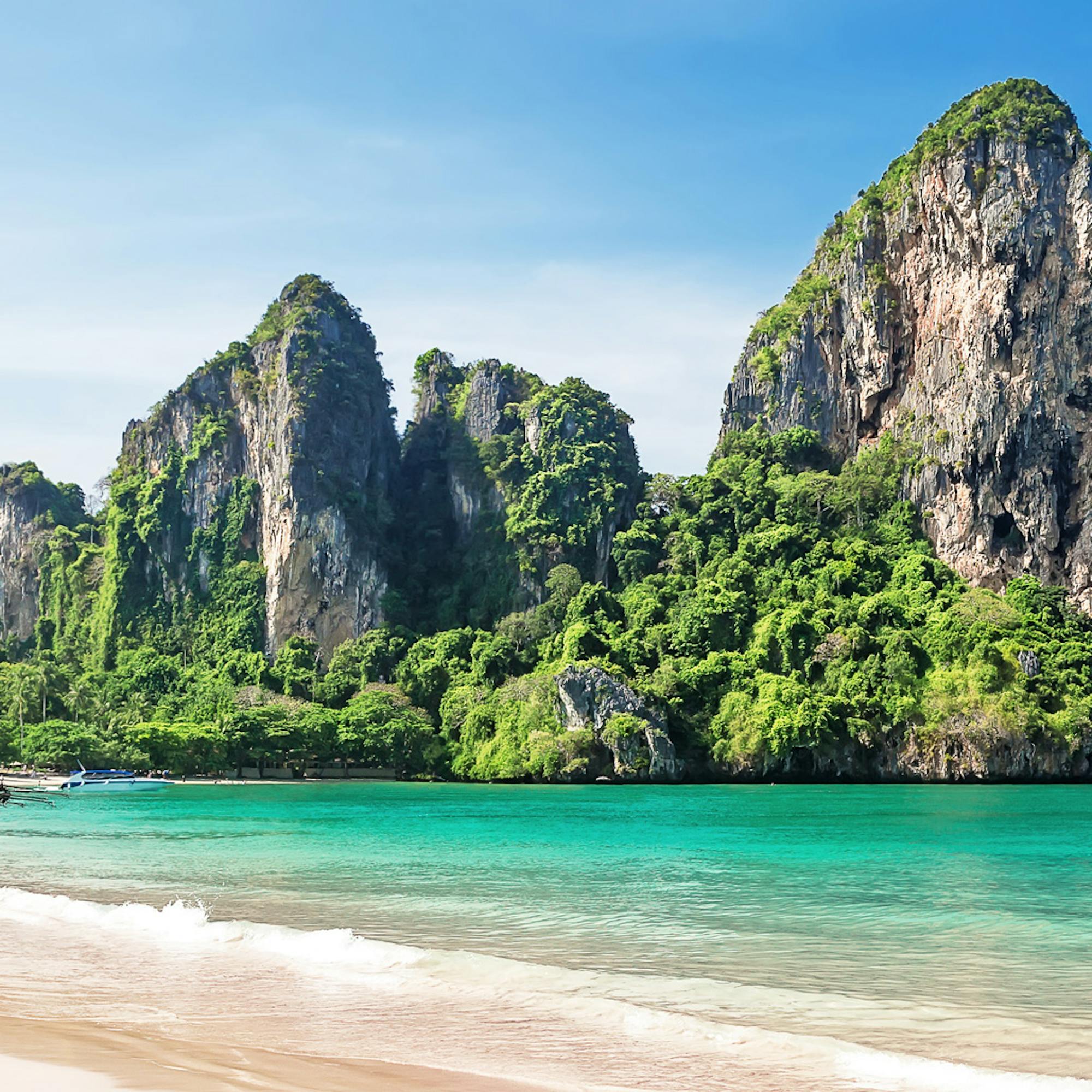 Limestone karst cliffs rise above Railay Beach, where pale sand meets clear turquoise water in bright sun.