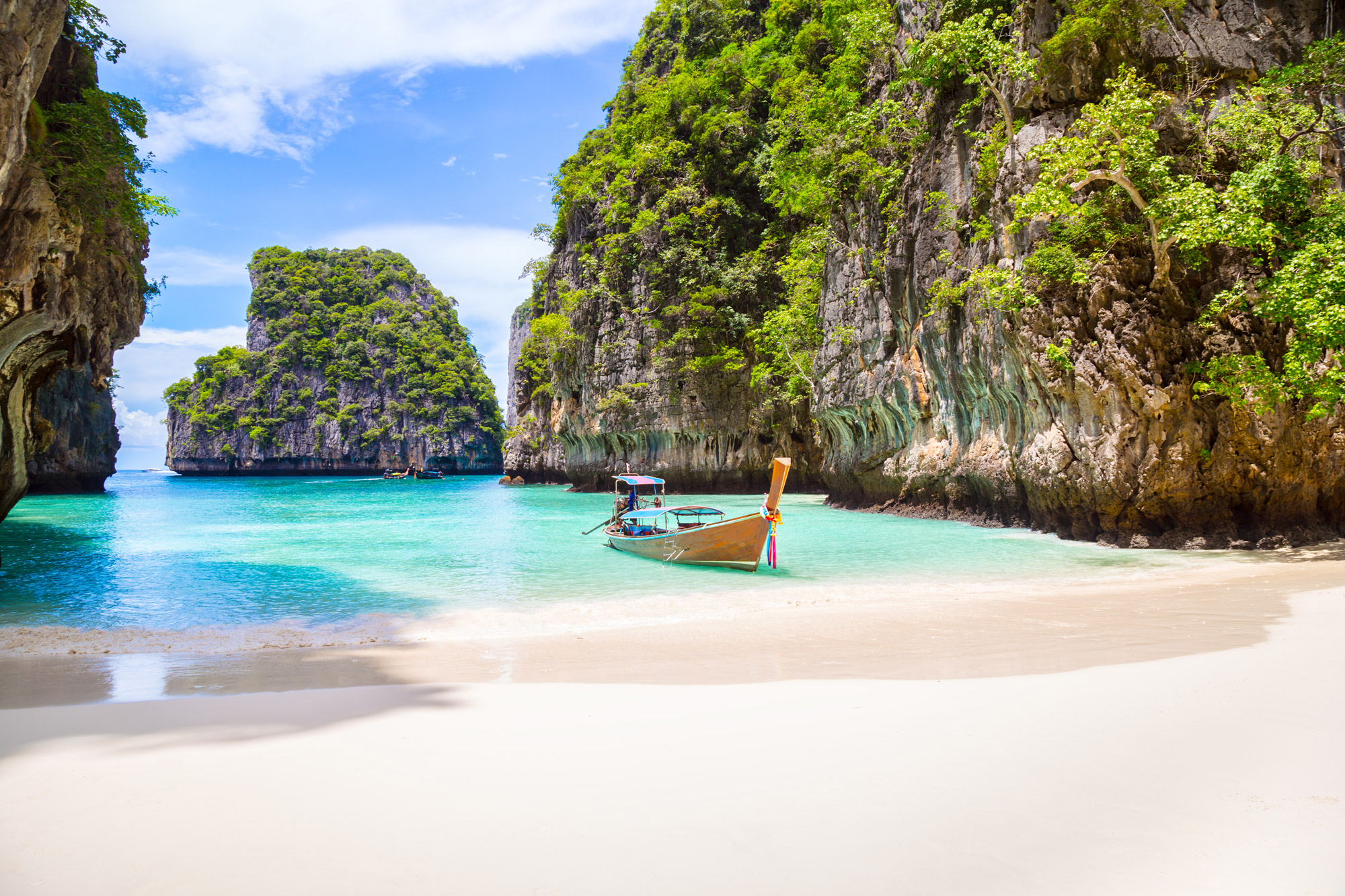 A traditional longtail boat floats in shallow blue water near Phuket’s limestone cliffs and a quiet sandy cove.