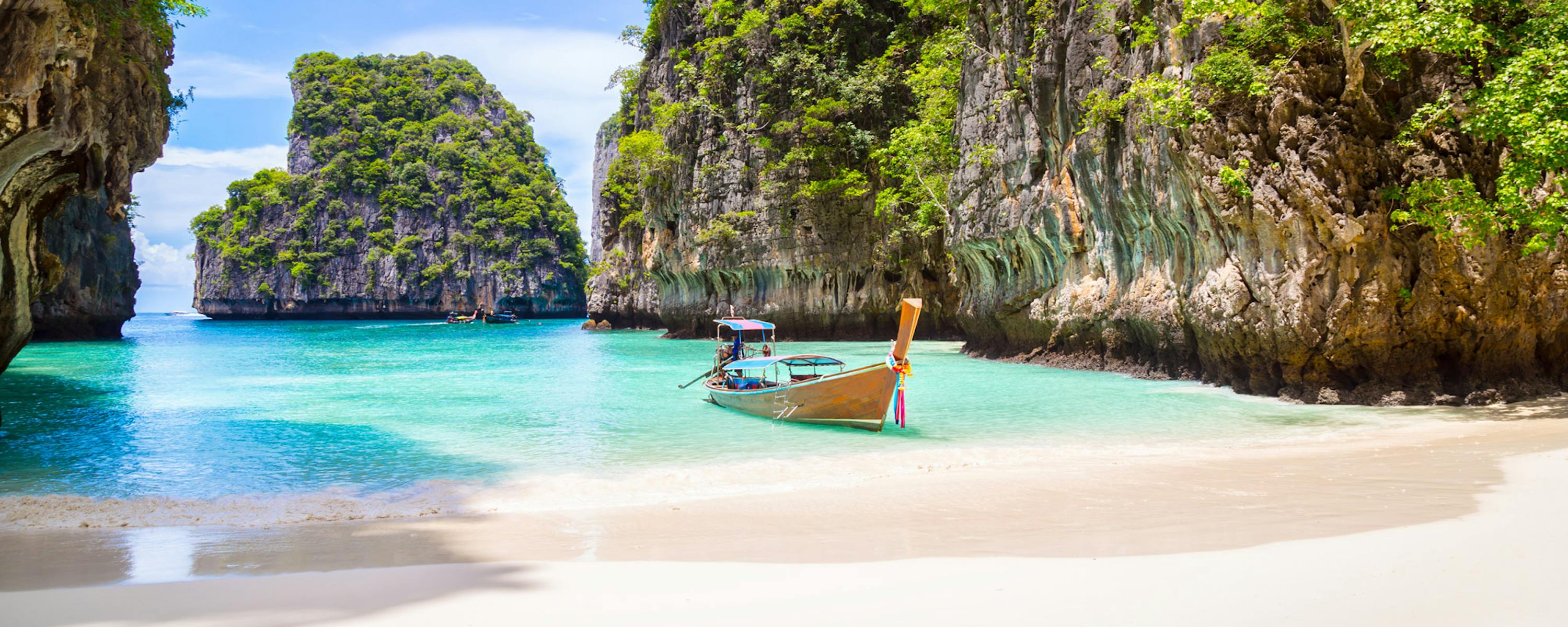 A traditional longtail boat floats in shallow blue water near Phuket’s limestone cliffs and a quiet sandy cove.