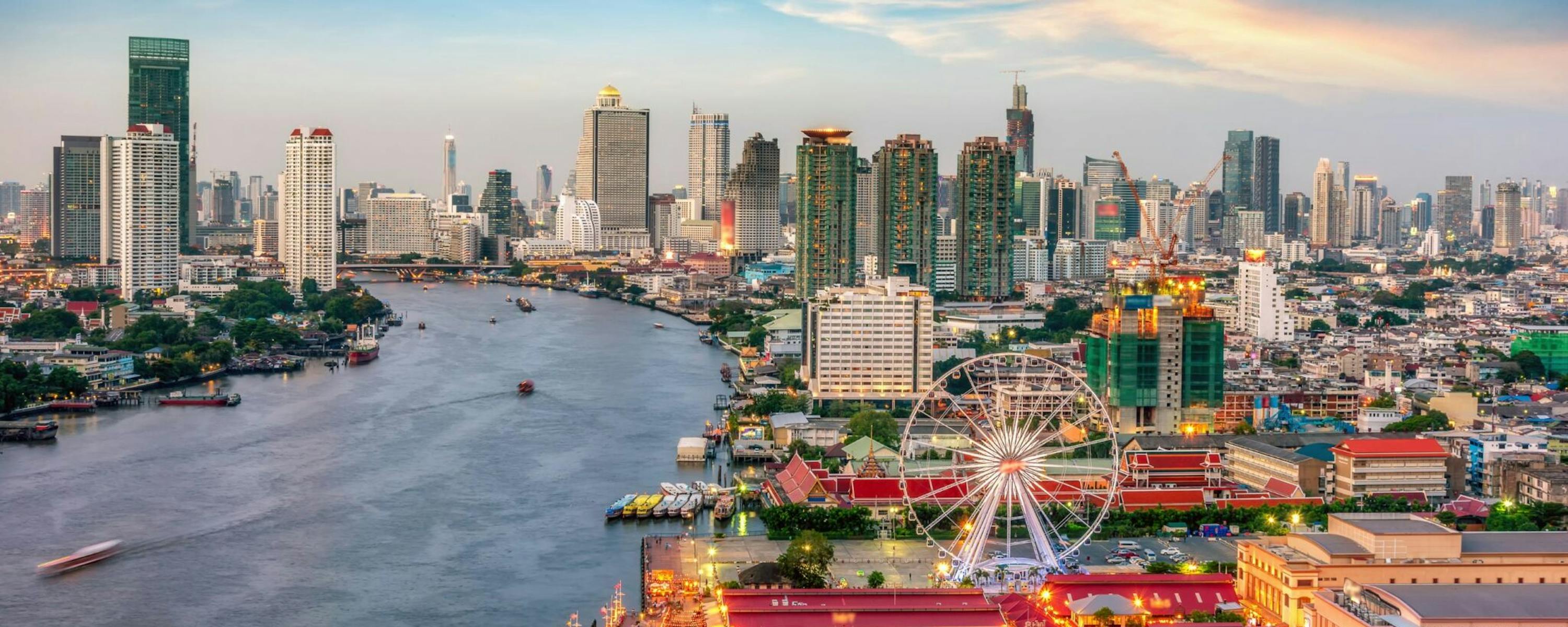 Bangkok’s skyline lines the Chao Phraya River at dusk, with temple rooftops in the foreground and boats below.