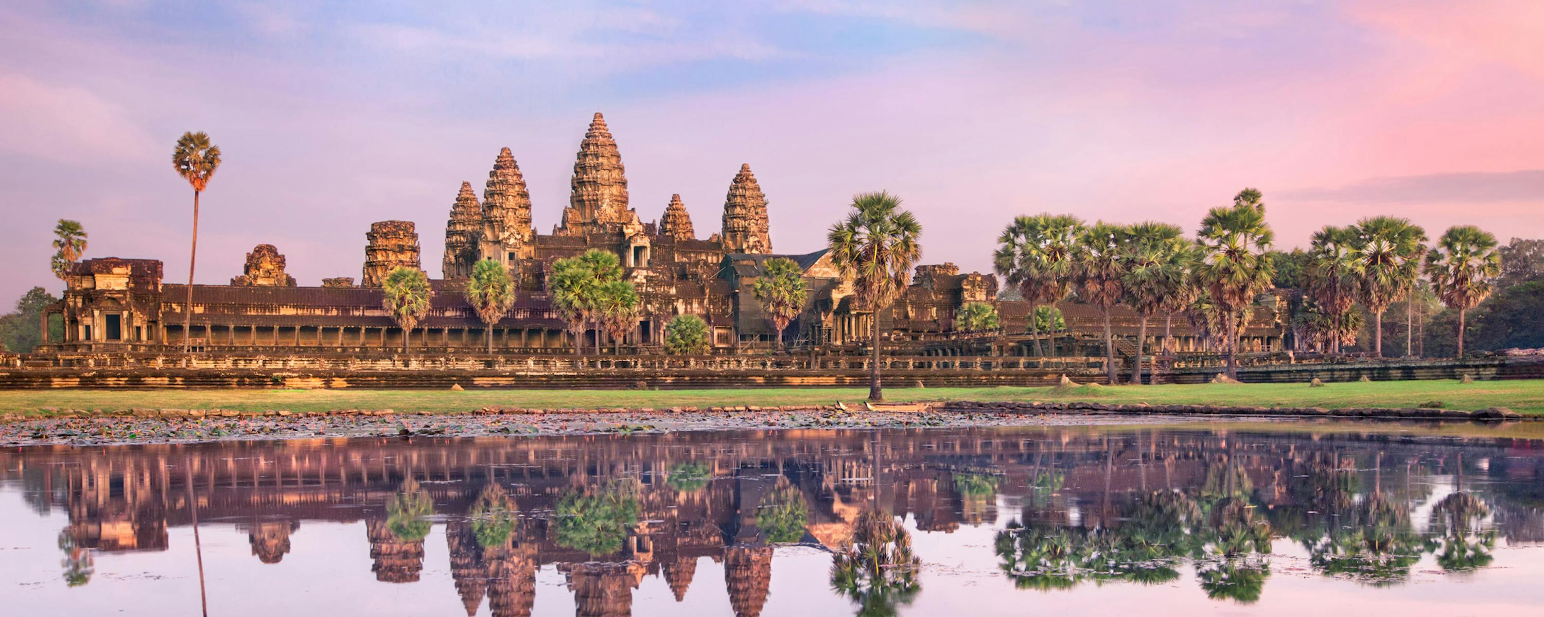 Angkor Wat silhouettes against a pink sunrise, mirrored in still water as palm trees frame the temple towers.