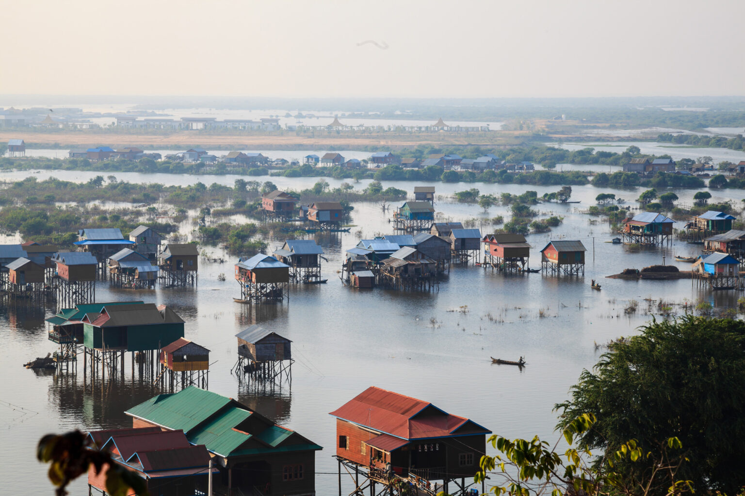 Stilt houses and colorful rooftops sit above floodwater, with small boats weaving through a broad lakeside community.