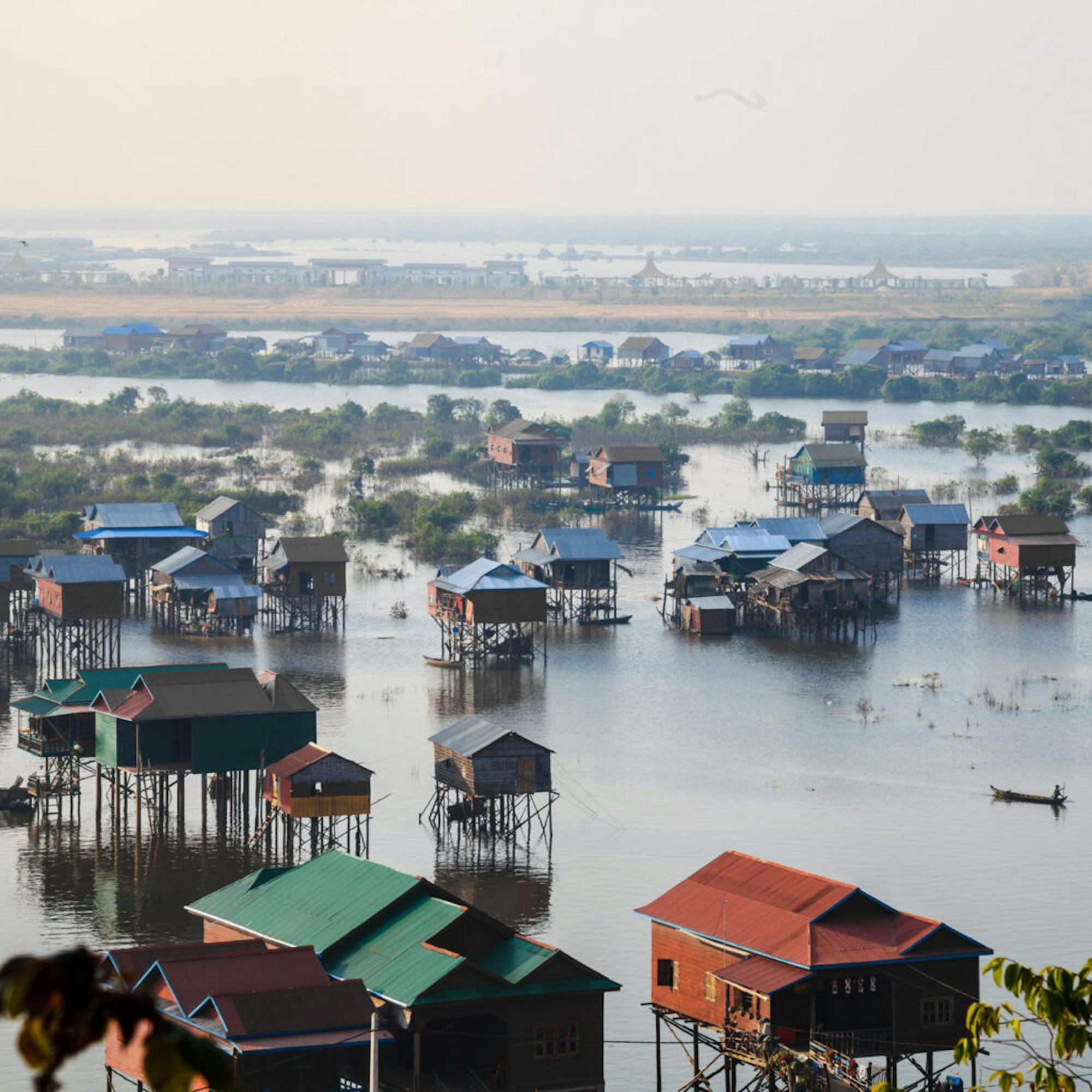 Stilt houses and colorful rooftops sit above floodwater, with small boats weaving through a broad lakeside community.