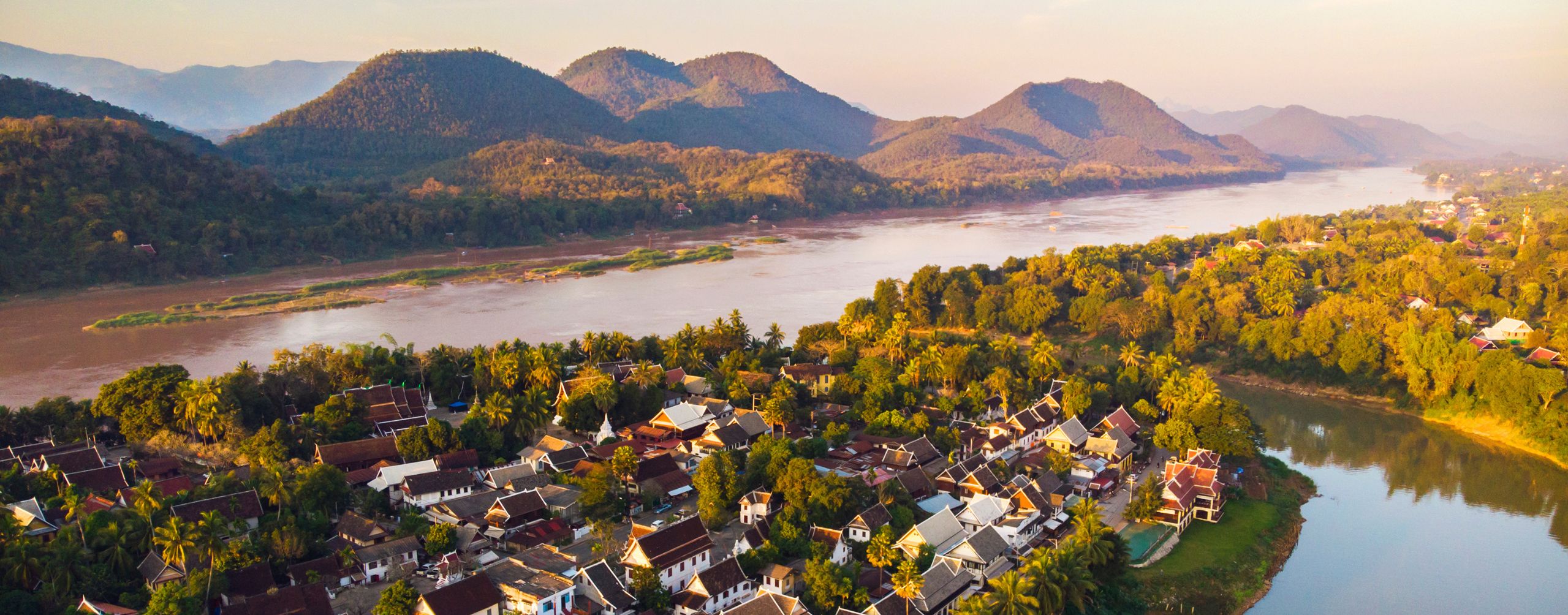 Luang Prabang sits beside a winding river, with sunlit trees, clustered rooftops, and mountains beyond the valley.