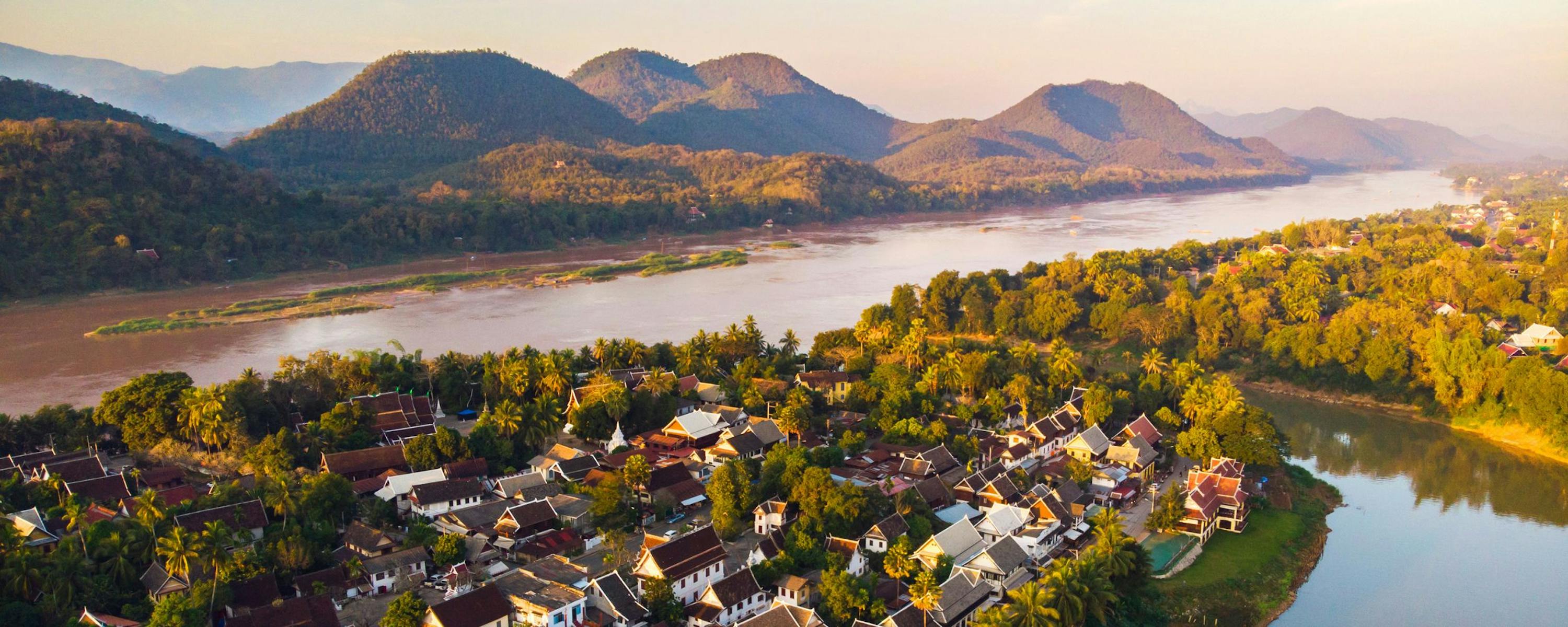 Luang Prabang sits beside a winding river, with sunlit trees, clustered rooftops, and mountains beyond the valley.