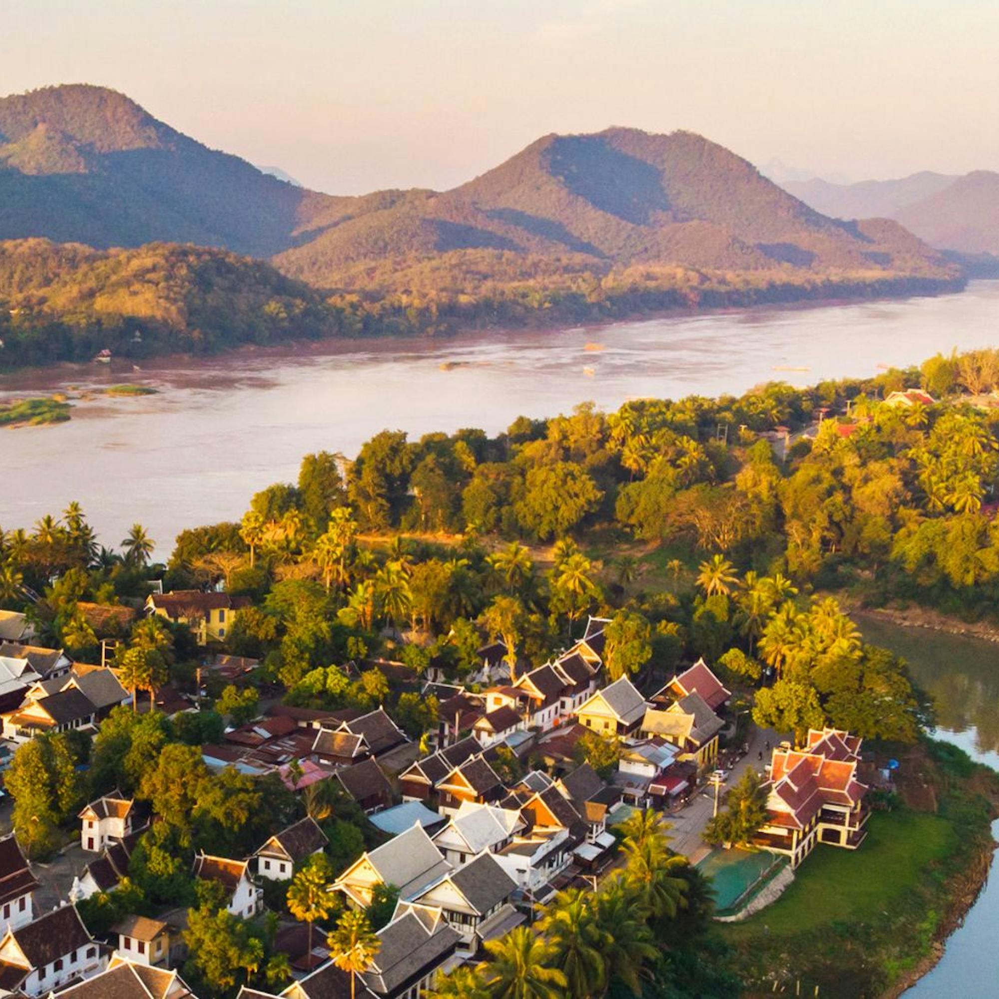 Luang Prabang sits beside a winding river, with sunlit trees, clustered rooftops, and mountains beyond the valley.