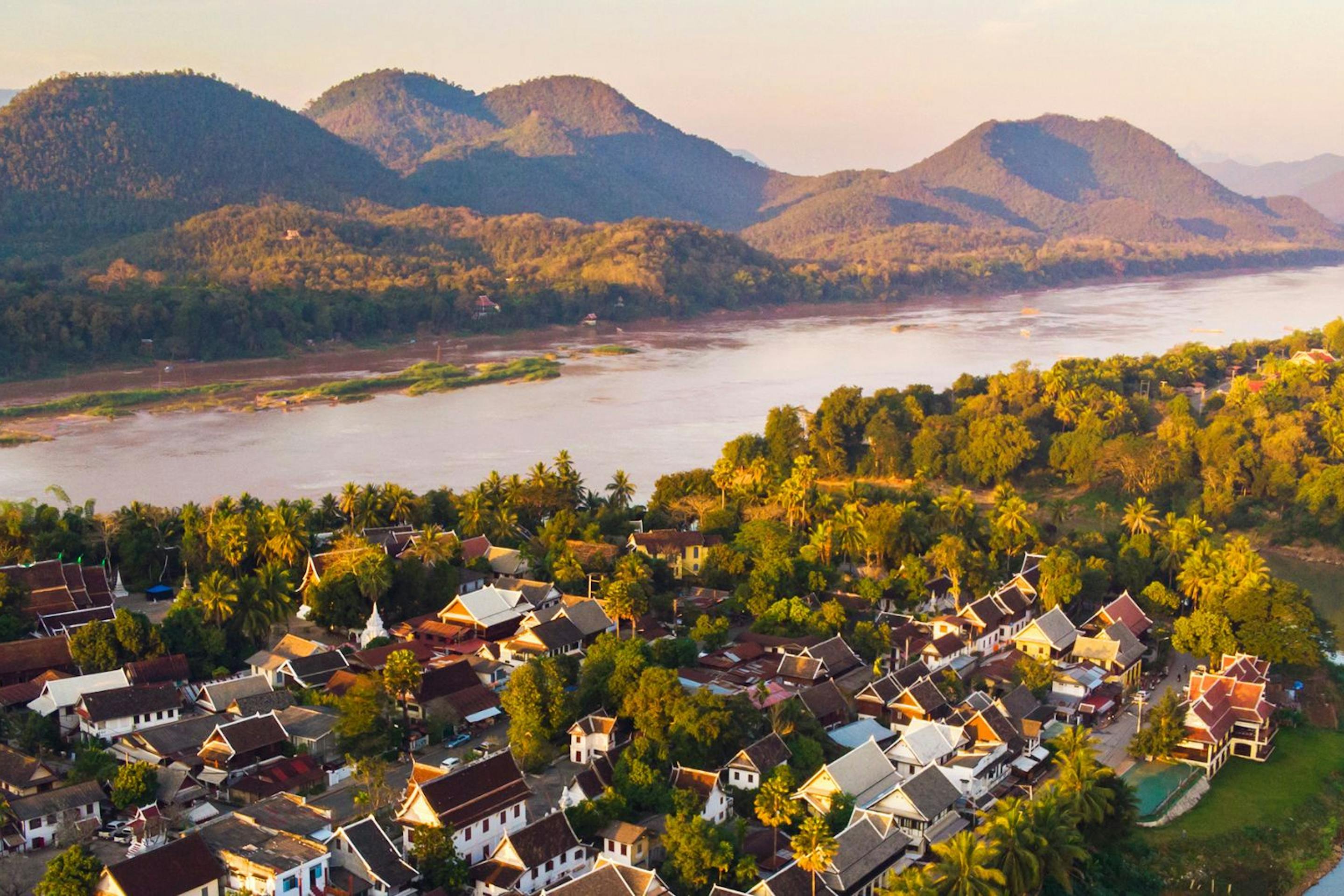 Luang Prabang sits beside a winding river, with sunlit trees, clustered rooftops, and mountains beyond the valley.