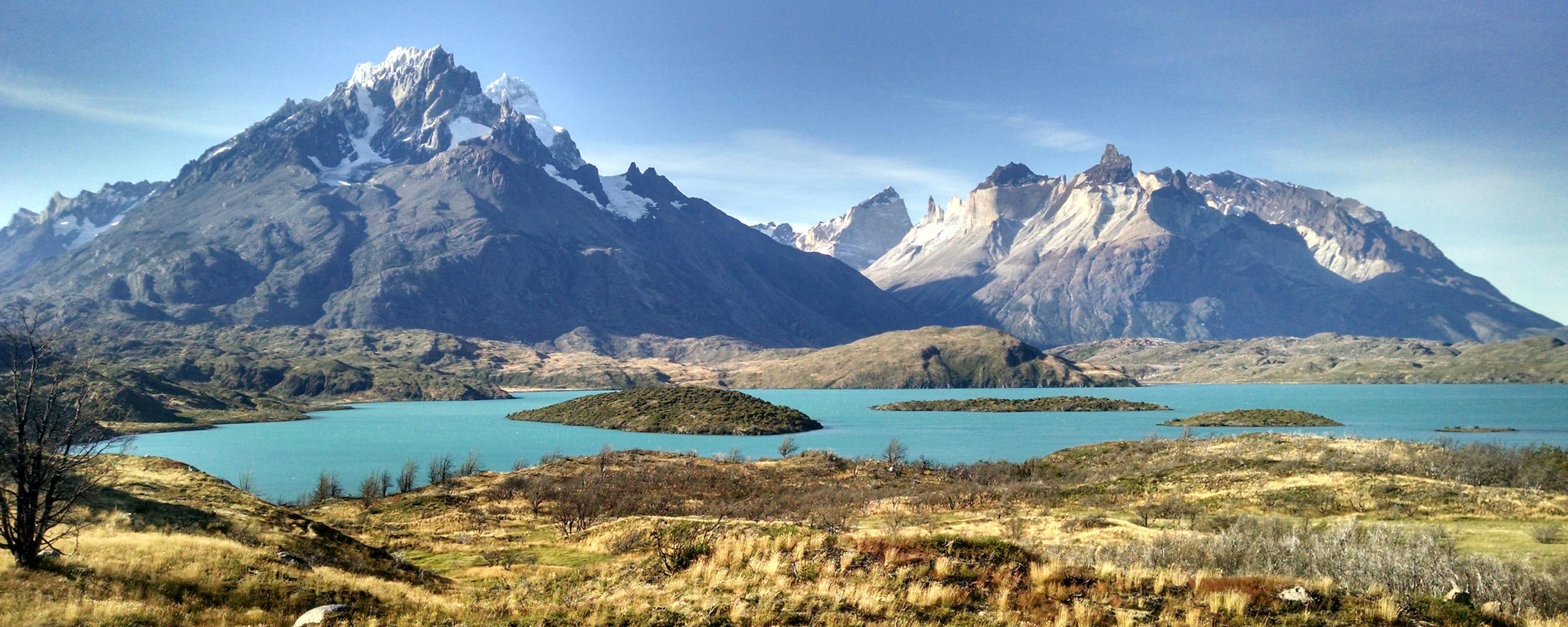 A turquoise lake winds through golden grasslands toward jagged, snowcapped peaks beneath a darkening sky.