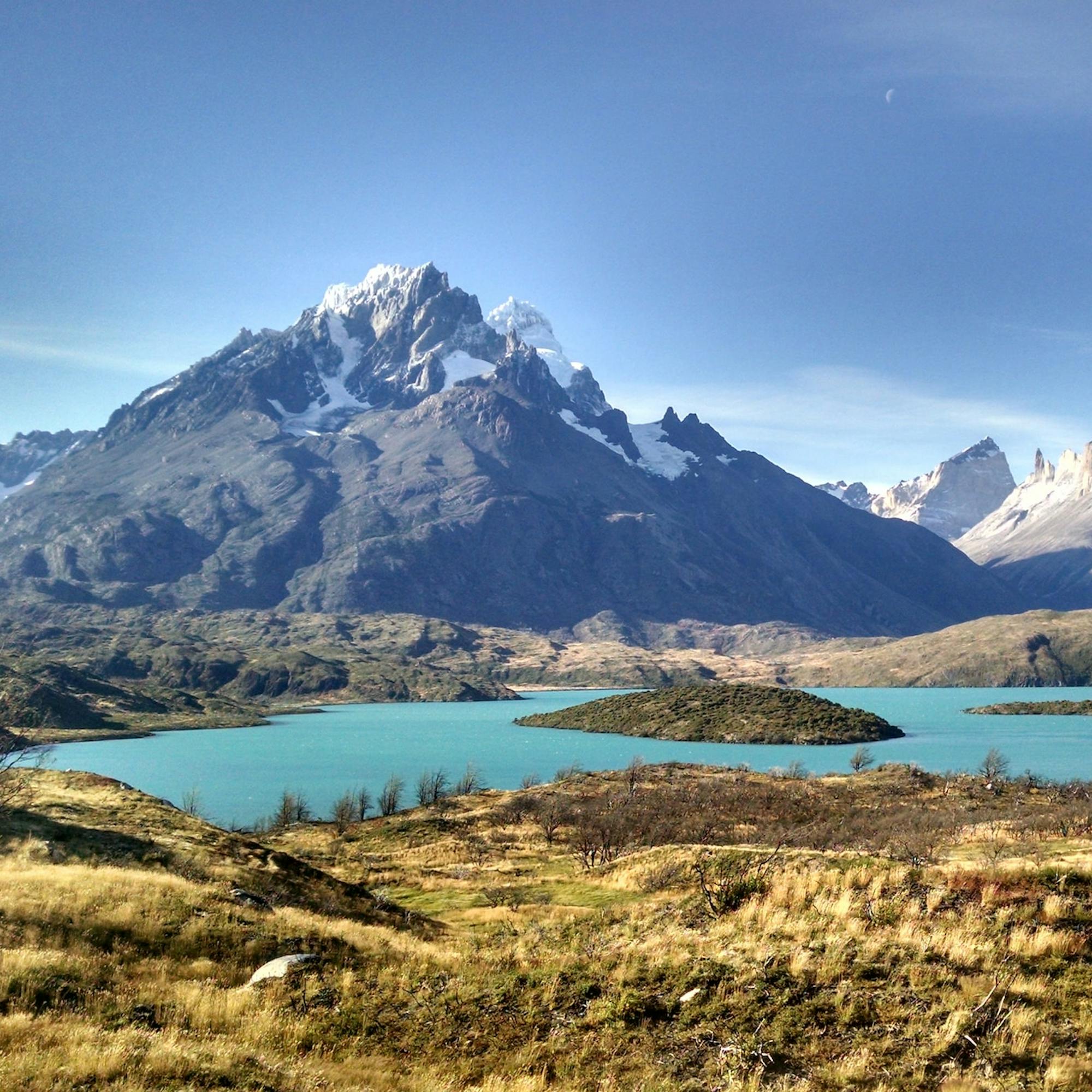 A turquoise lake winds through golden grasslands toward jagged, snowcapped peaks beneath a darkening sky.