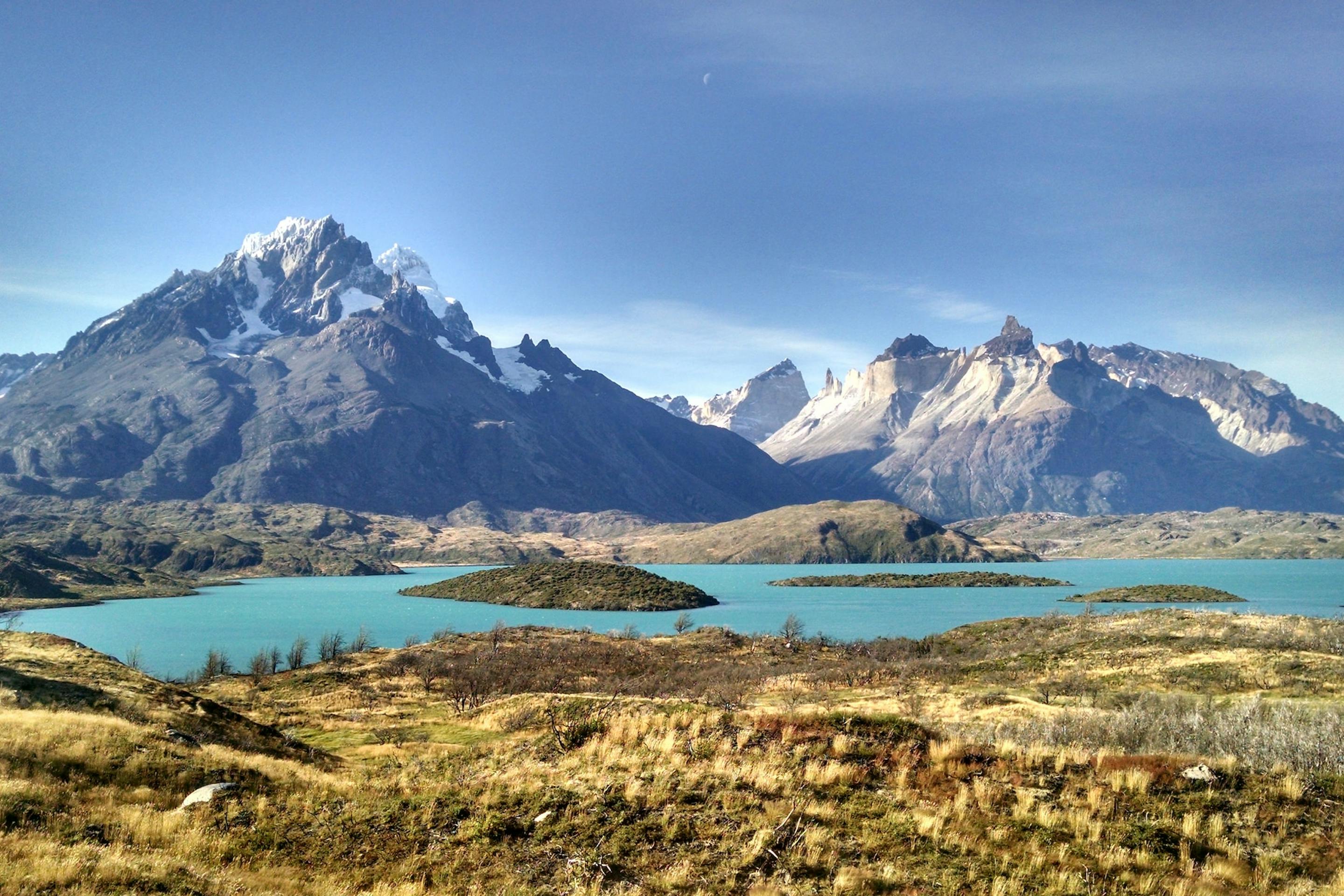 A turquoise lake winds through golden grasslands toward jagged, snowcapped peaks beneath a darkening sky.