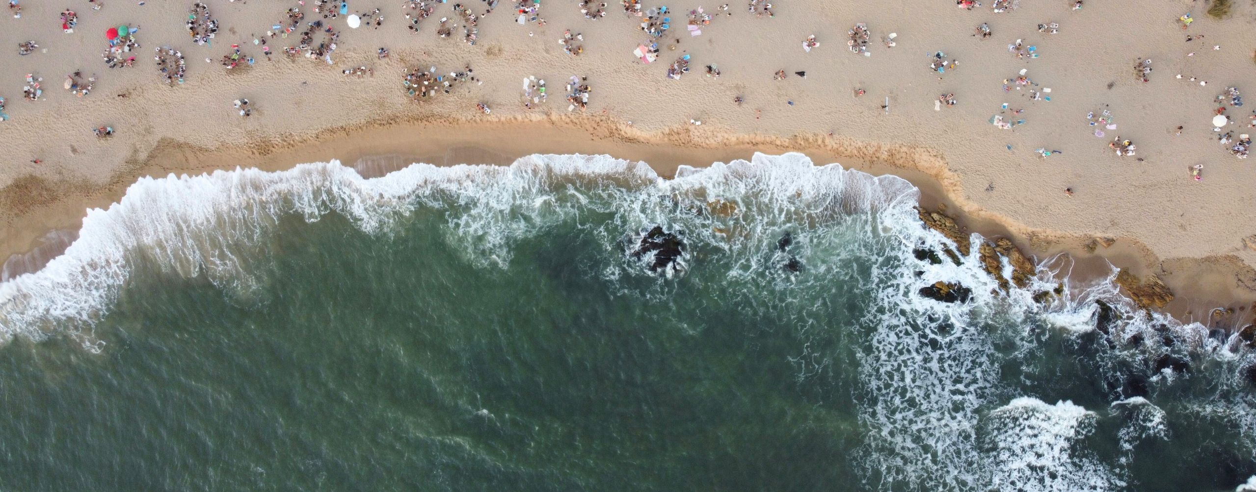 Aerial view of surf curling onto a sandy beach dotted with swimmers and umbrellas, leaving foam along the shore.