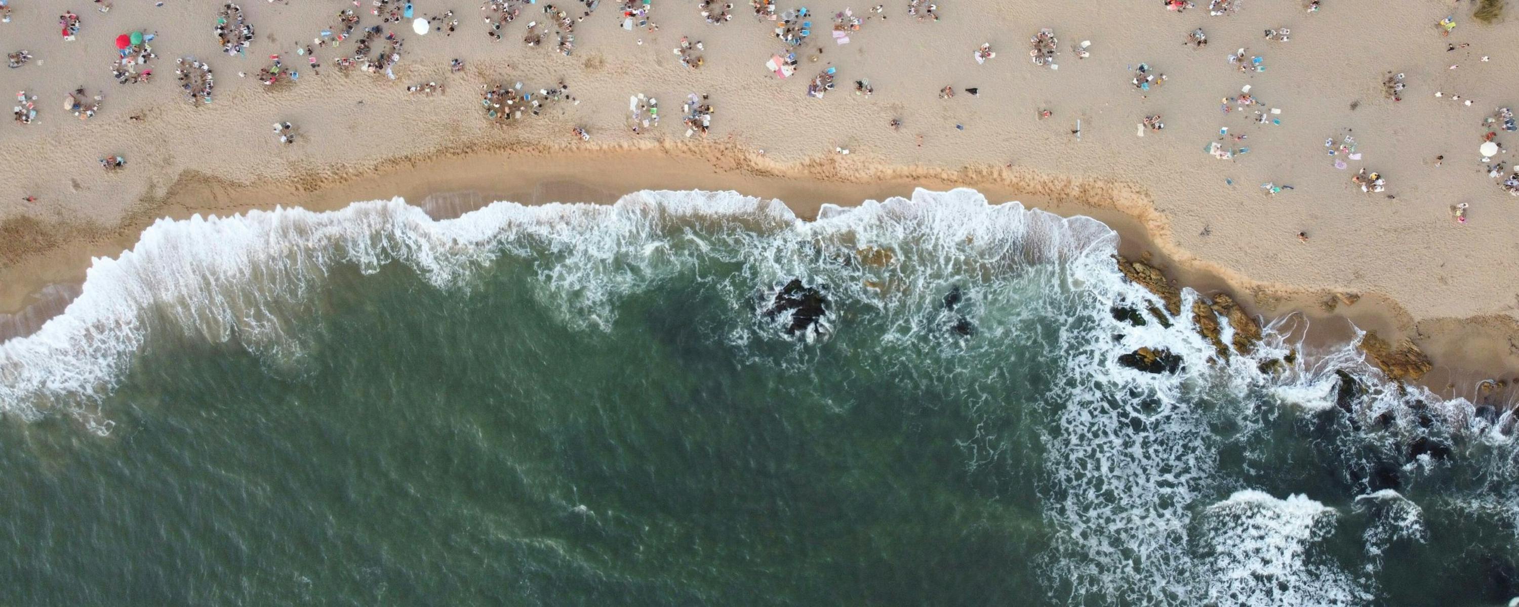 Aerial view of surf curling onto a sandy beach dotted with swimmers and umbrellas, leaving foam along the shore.
