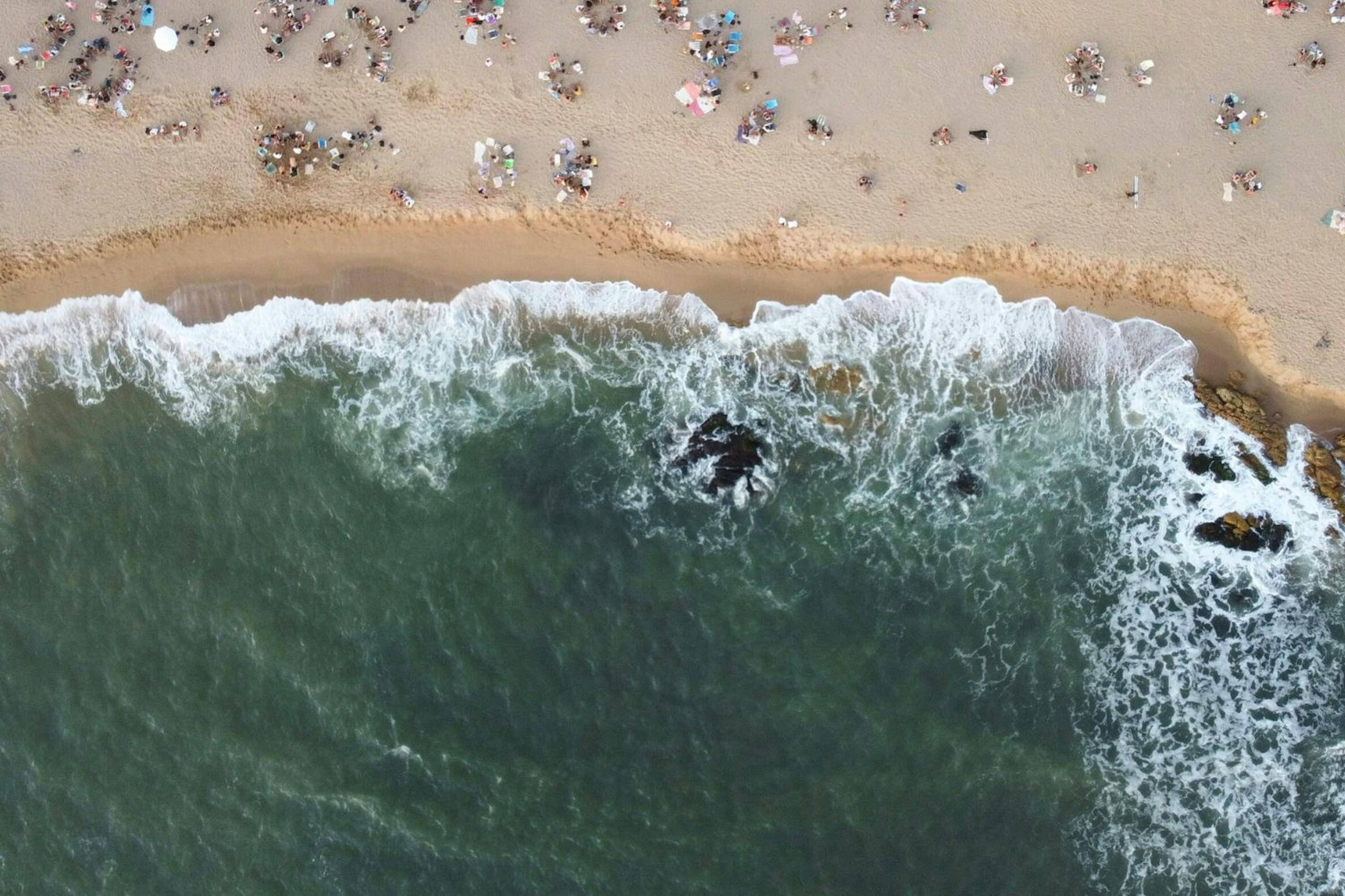 Aerial view of surf curling onto a sandy beach dotted with swimmers and umbrellas, leaving foam along the shore.