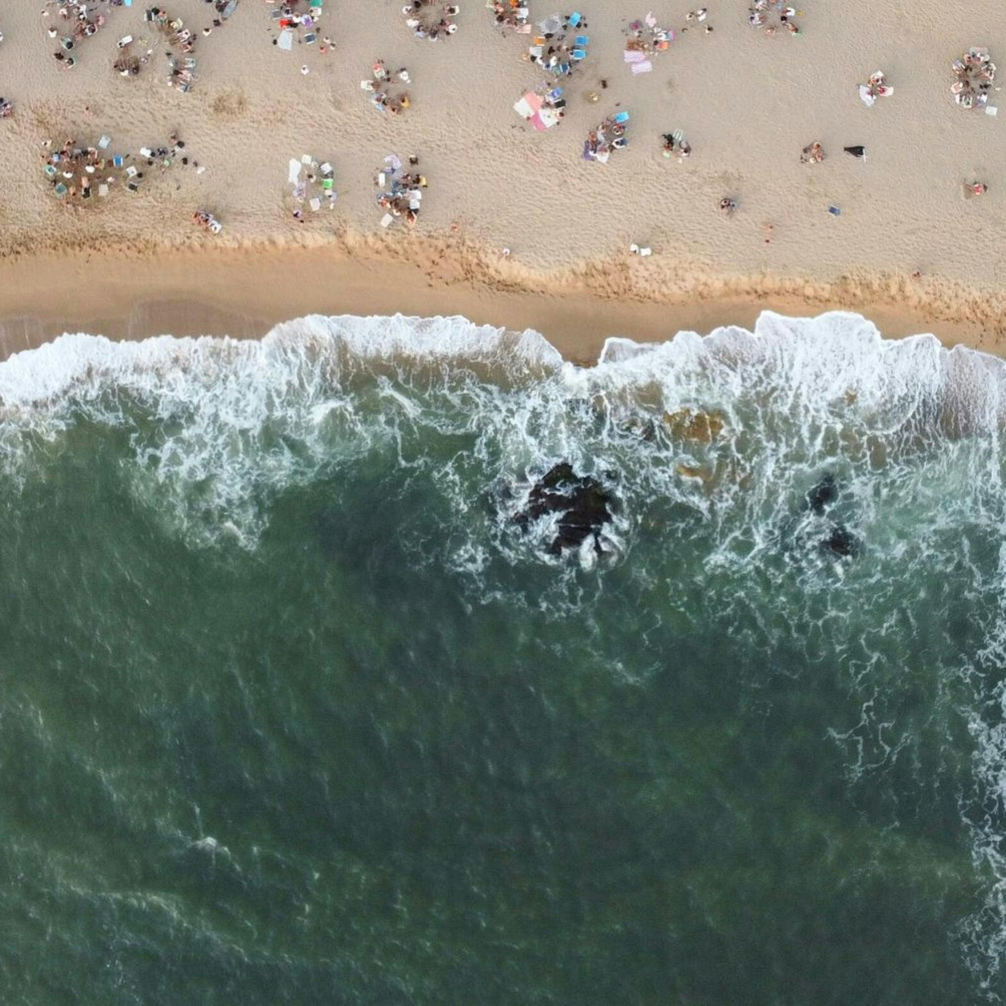 Aerial view of surf curling onto a sandy beach dotted with swimmers and umbrellas, leaving foam along the shore.