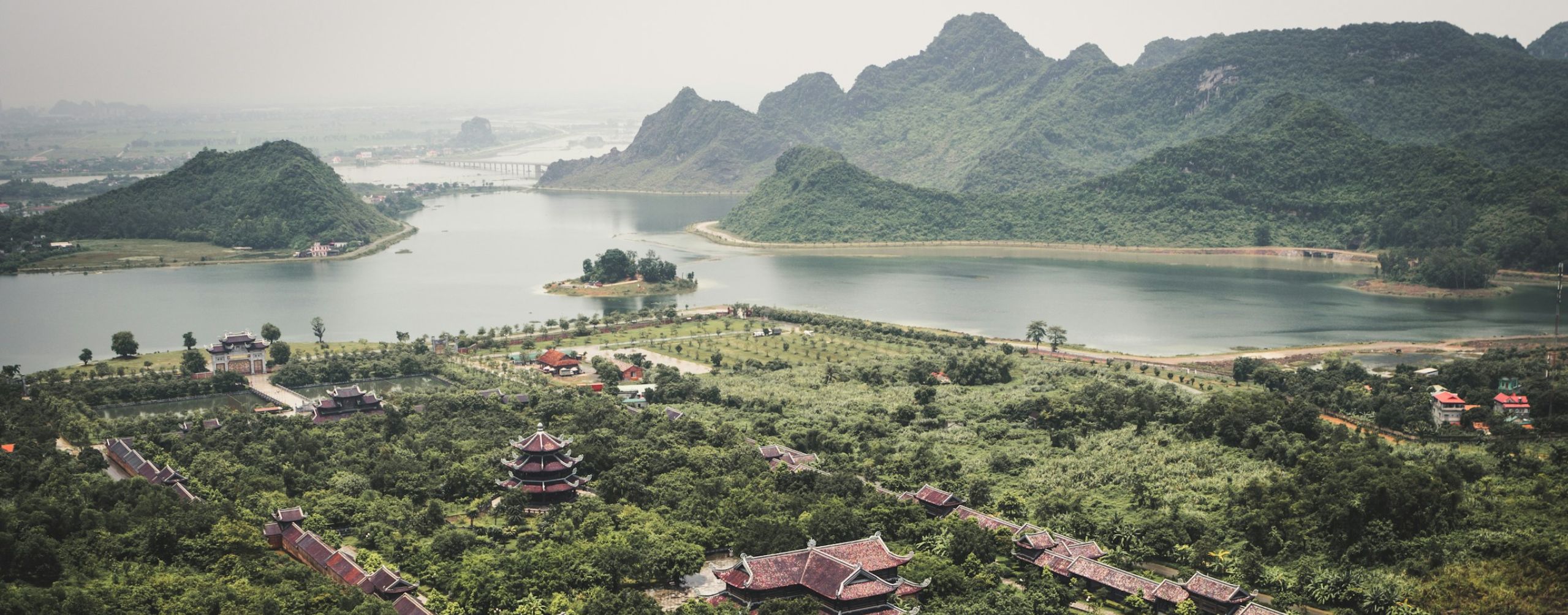 Green karst peaks rise around a winding lake and village rooftops, with boats moving slowly across hazy water.