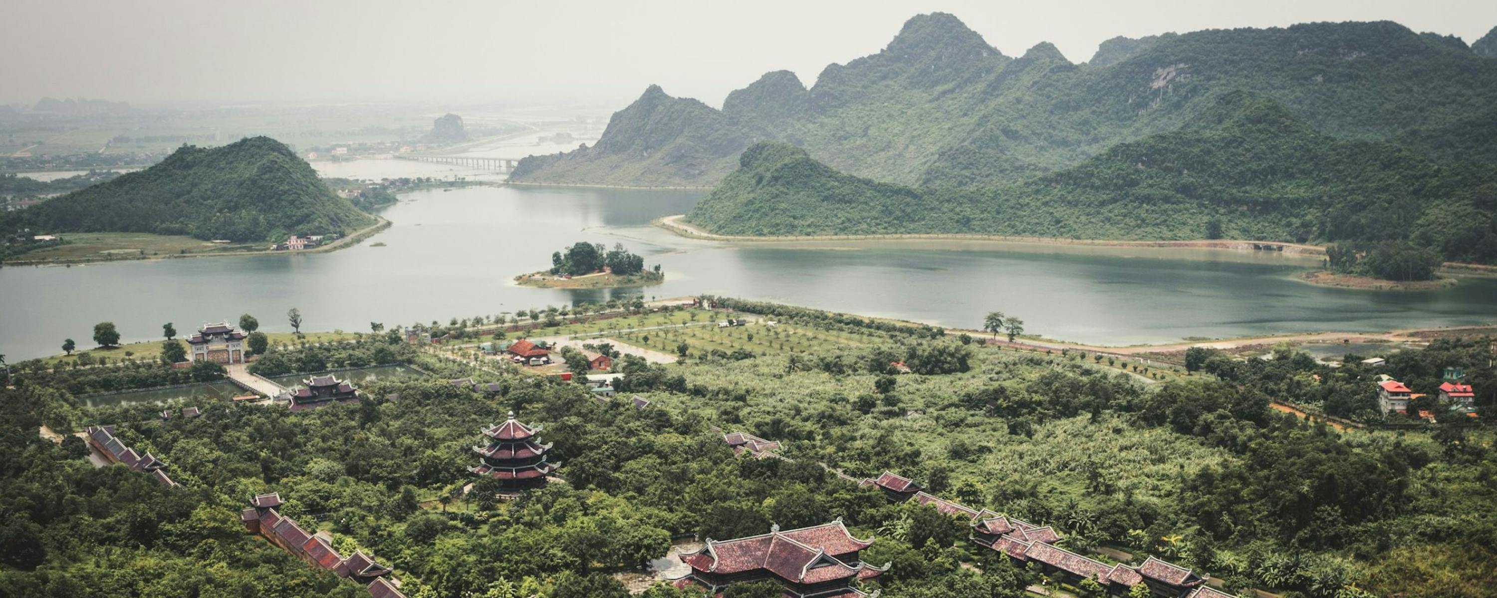 Green karst peaks rise around a winding lake and village rooftops, with boats moving slowly across hazy water.