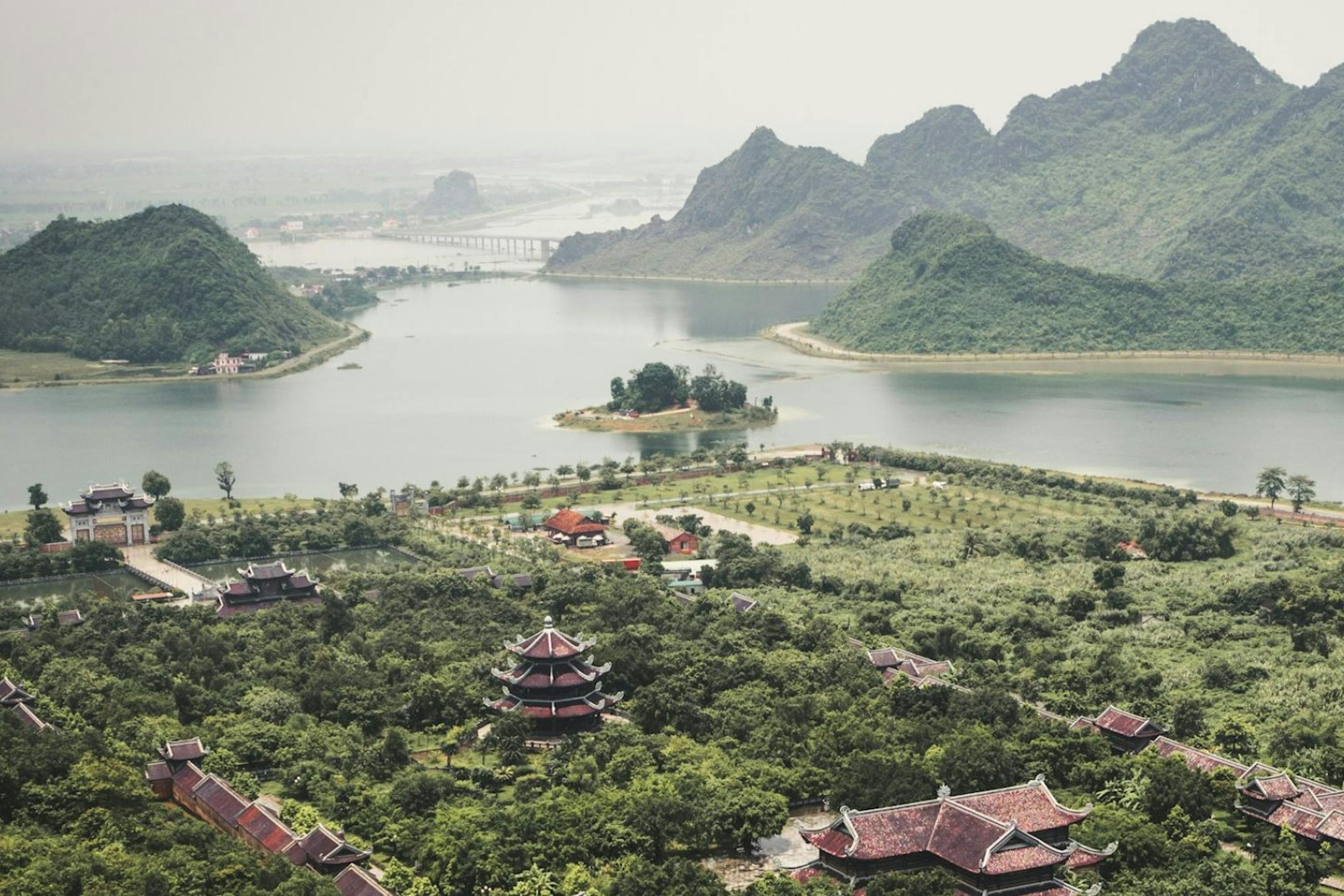 Green karst peaks rise around a winding lake and village rooftops, with boats moving slowly across hazy water.