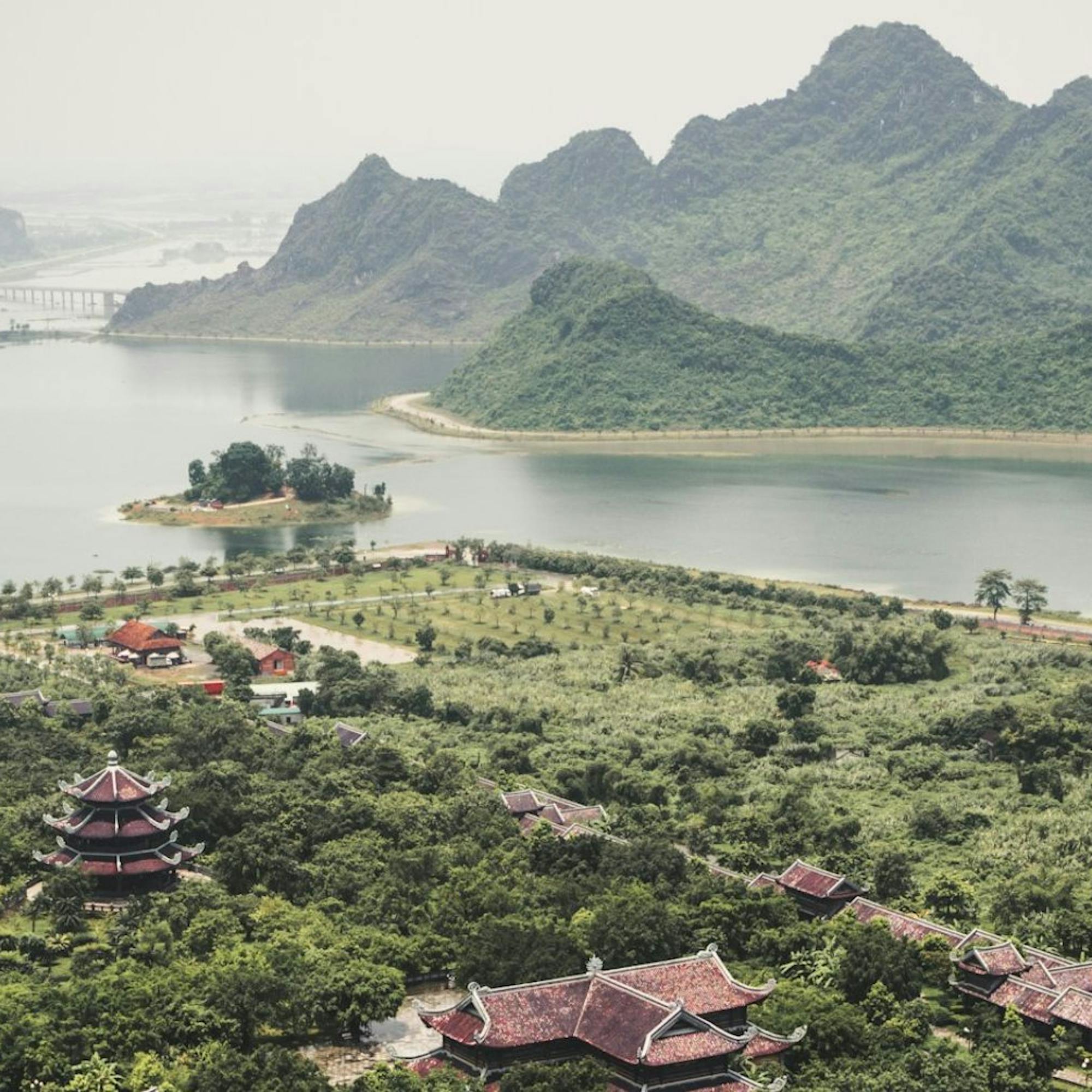 Green karst peaks rise around a winding lake and village rooftops, with boats moving slowly across hazy water.