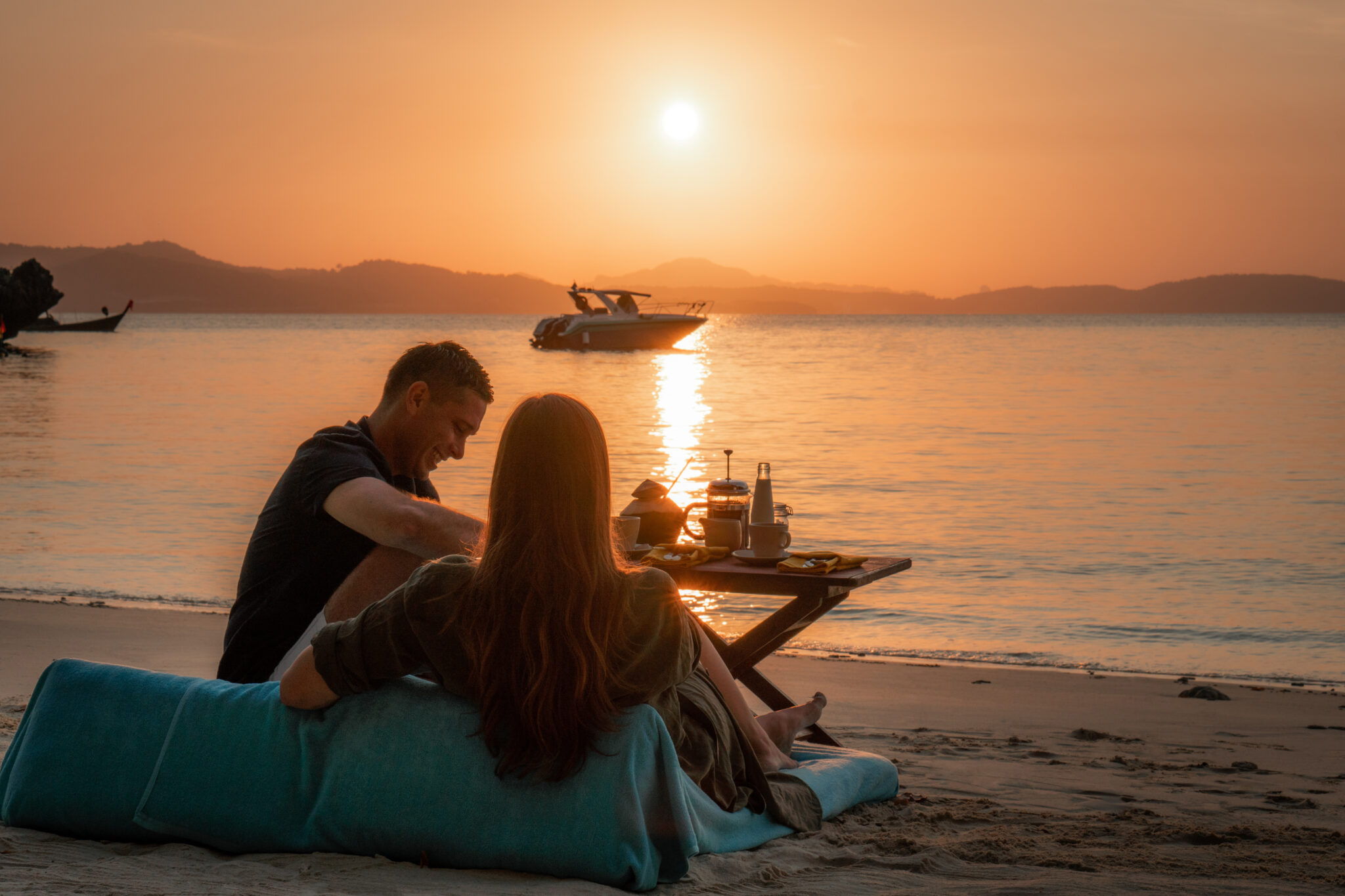 Couple sits on a beach beanbag at sunrise with a breakfast spread and longtail boats anchored on calm water.