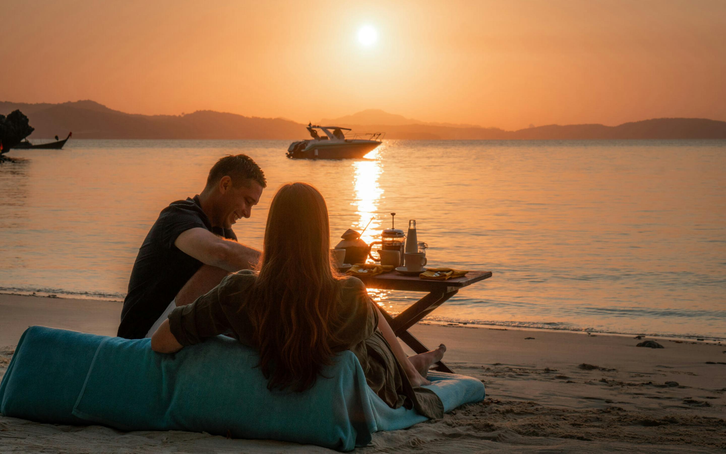 Couple sits on a beach beanbag at sunrise with a breakfast spread and longtail boats anchored on calm water.