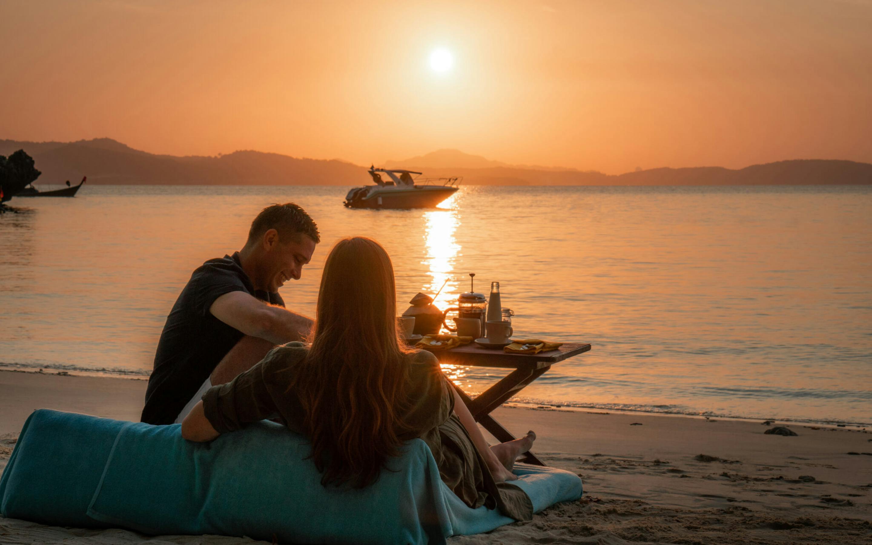 Couple sits on a beach beanbag at sunrise with a breakfast spread and longtail boats anchored on calm water.