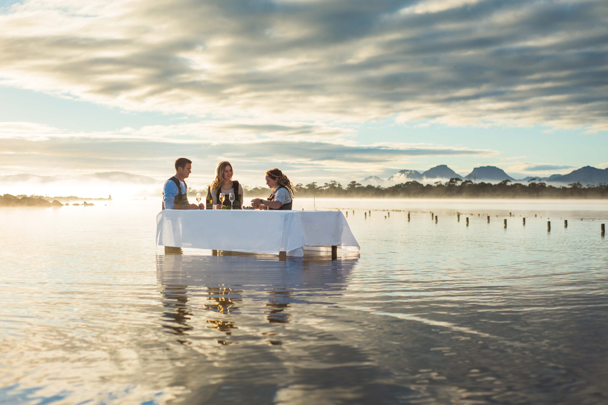 A small group sits around a table set in shallow water, shucking oysters as low waves ripple under a wide sky.