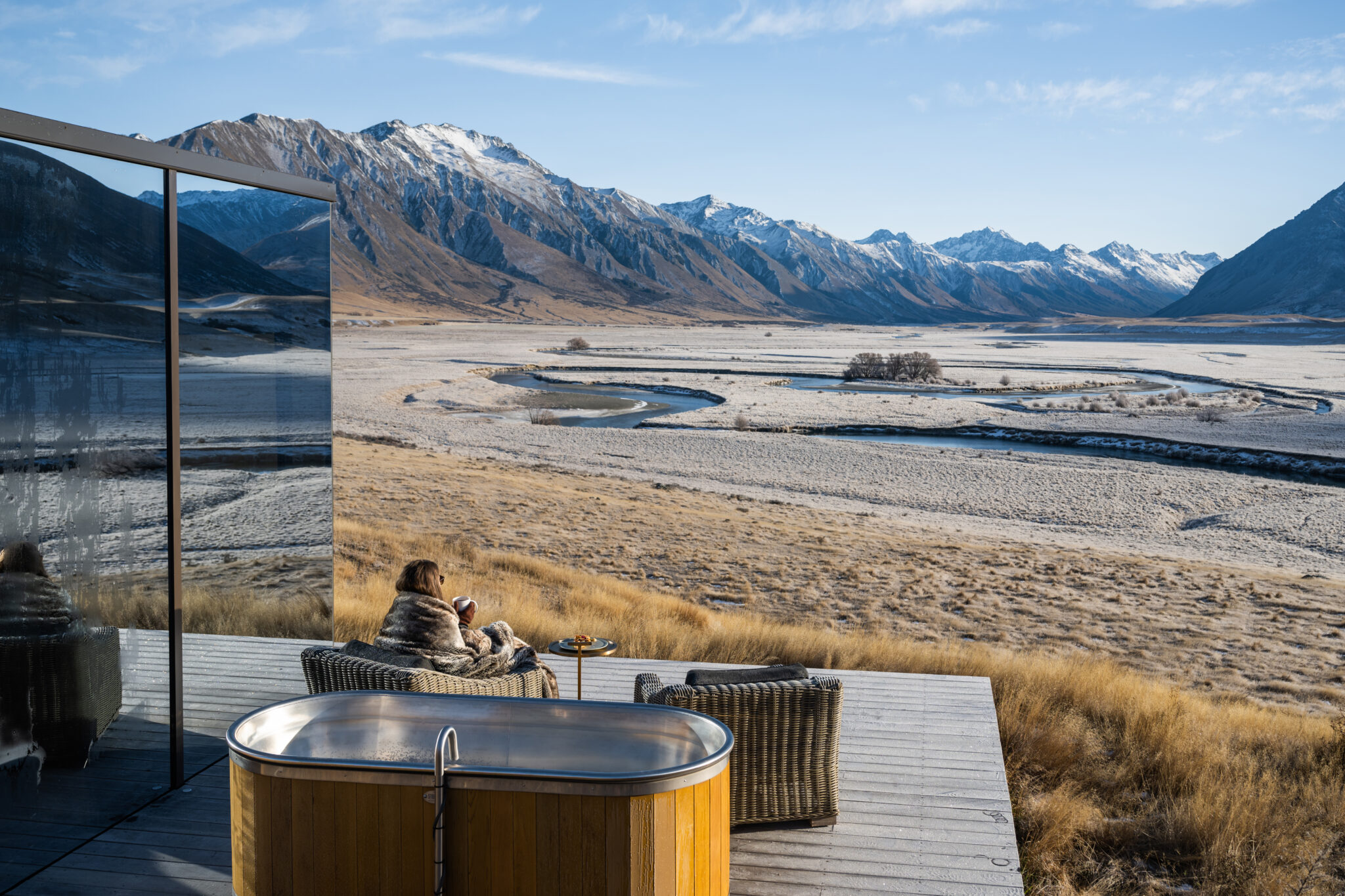 A round hot tub on a deck looks across a wide riverbed toward snow-dusted mountains under clear blue skies.
