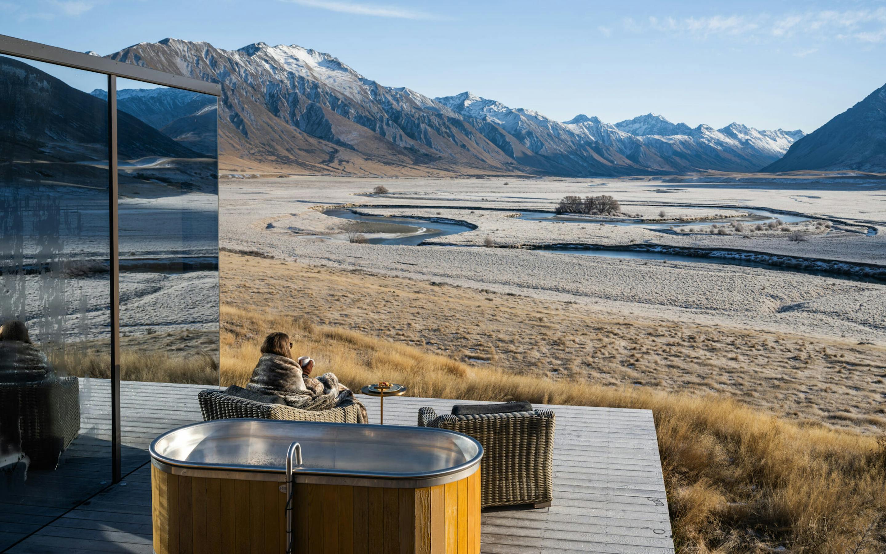 A round hot tub on a deck looks across a wide riverbed toward snow-dusted mountains under clear blue skies.