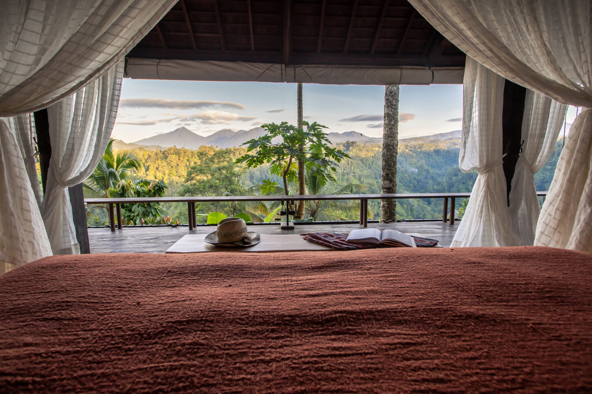 Canopied bed inside an open-air suite frames misty green valley views through wide glass doors and curtains.