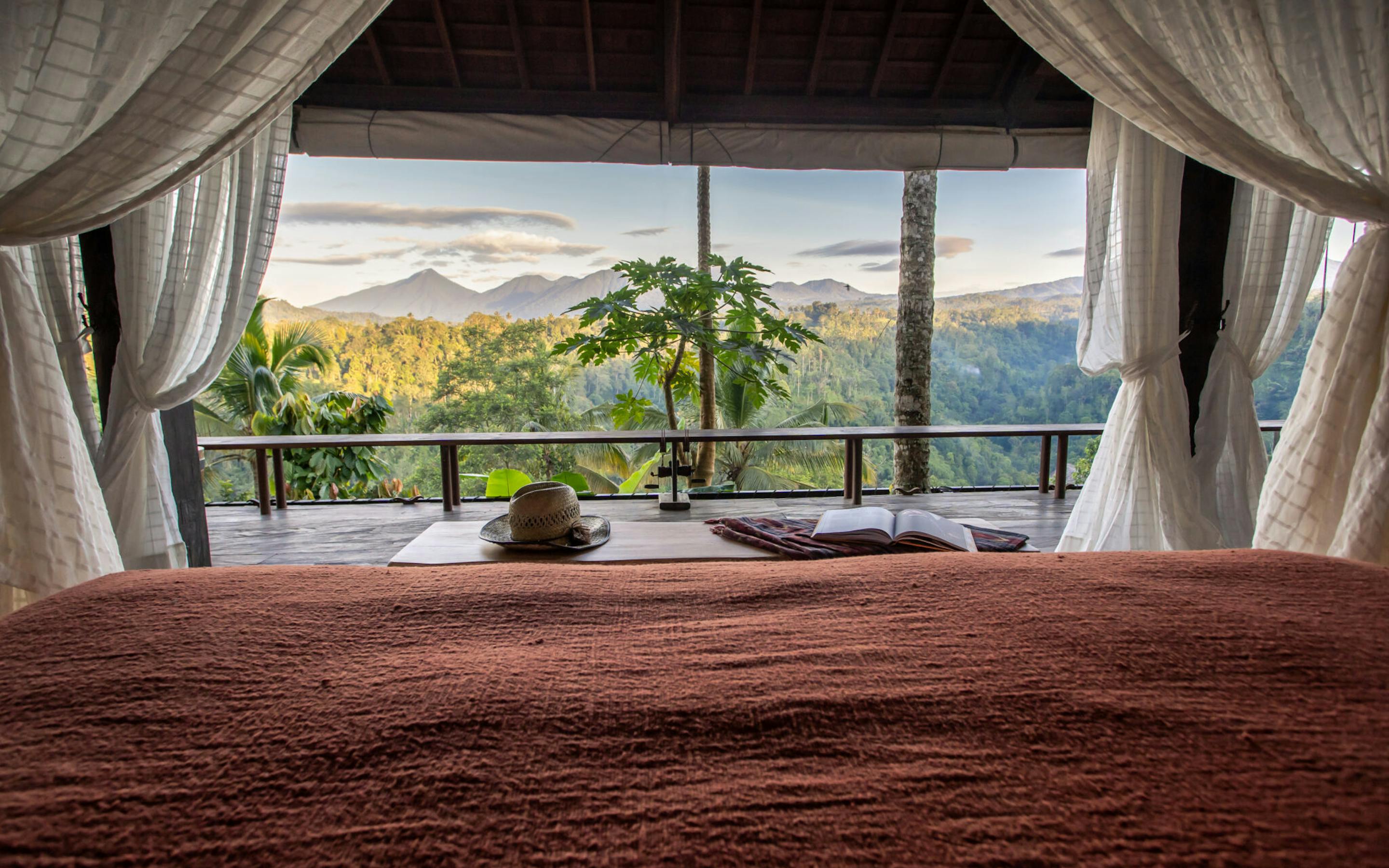 Canopied bed inside an open-air suite frames misty green valley views through wide glass doors and curtains.