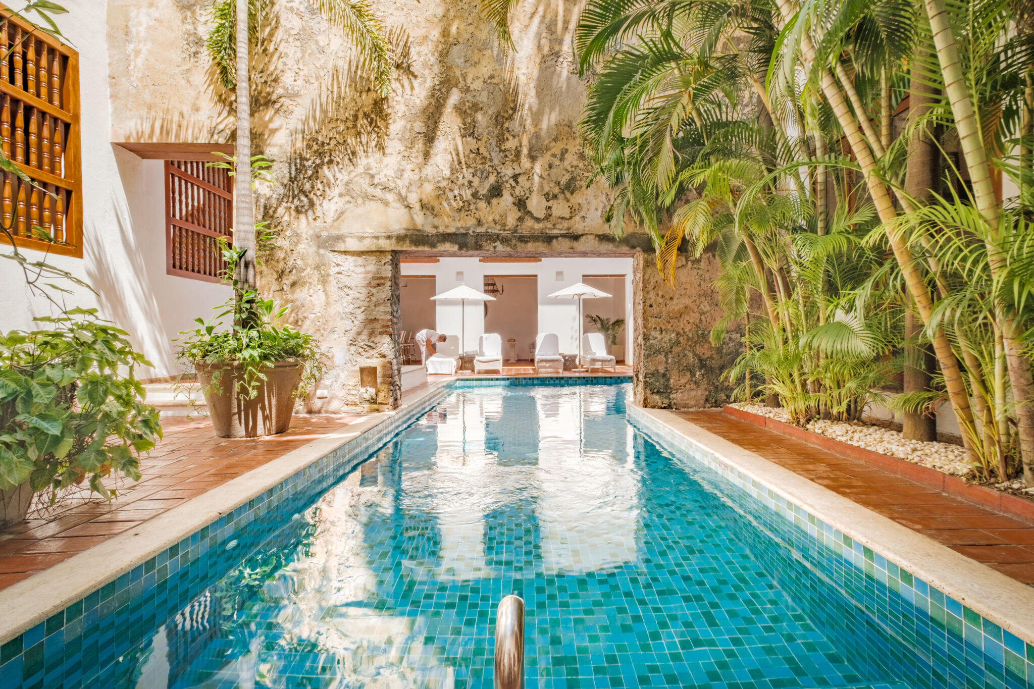 A long turquoise pool runs through a leafy courtyard, with white walls, arches, and sunlit tiles at the far end.