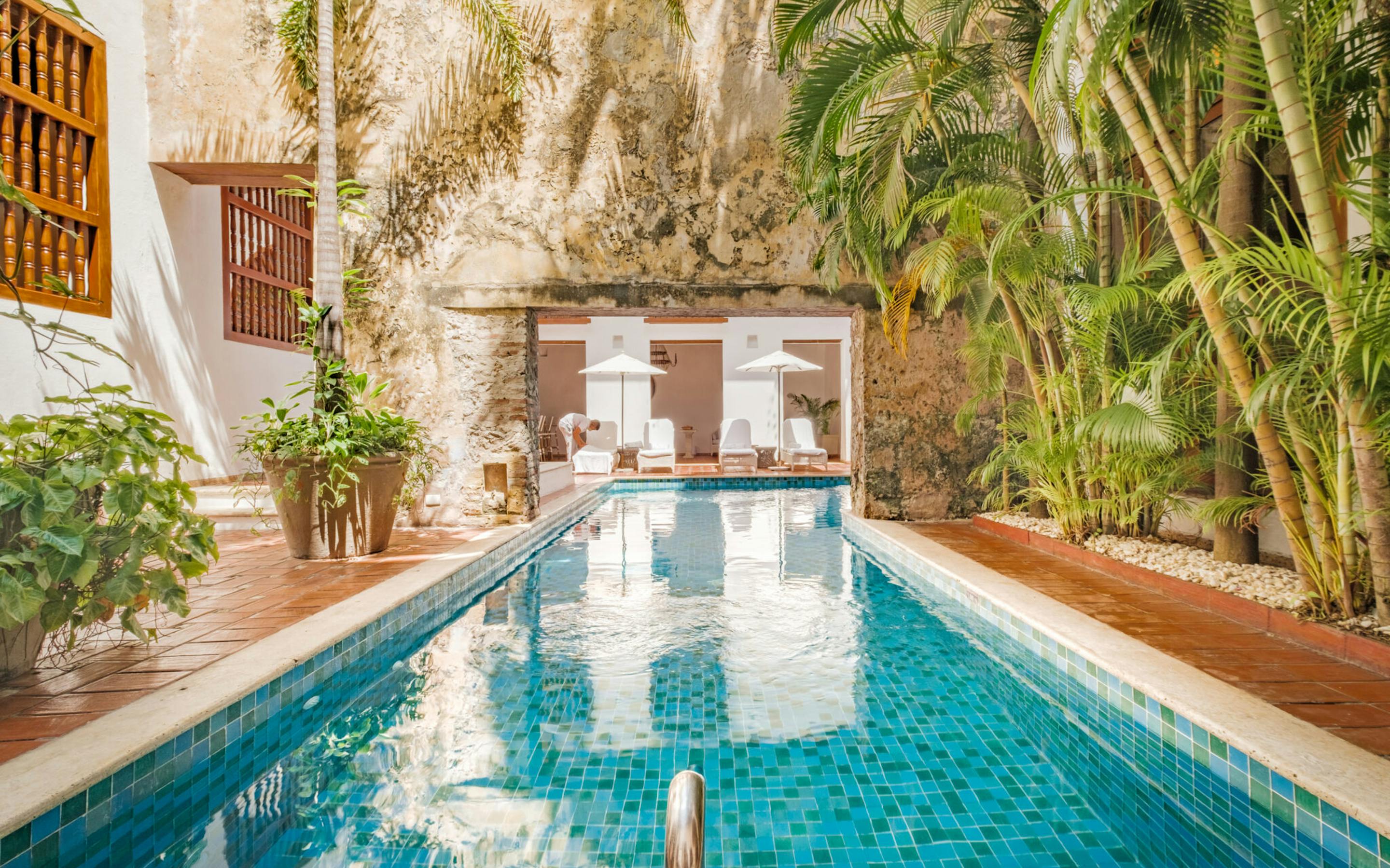 A long turquoise pool runs through a leafy courtyard, with white walls, arches, and sunlit tiles at the far end.