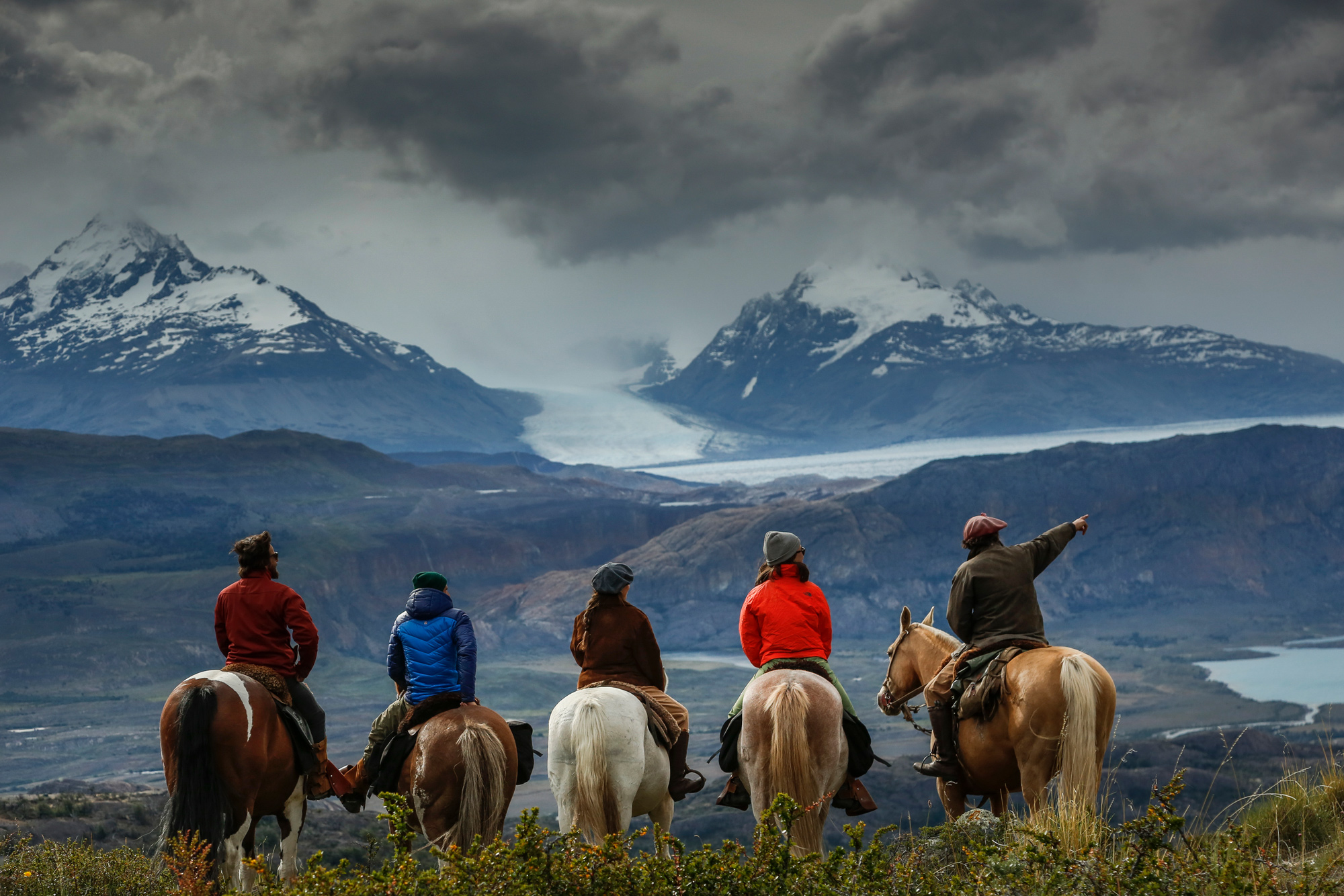 Riders on horseback cross open plains toward snow-dusted mountains beneath heavy clouds and misty ridgelines.