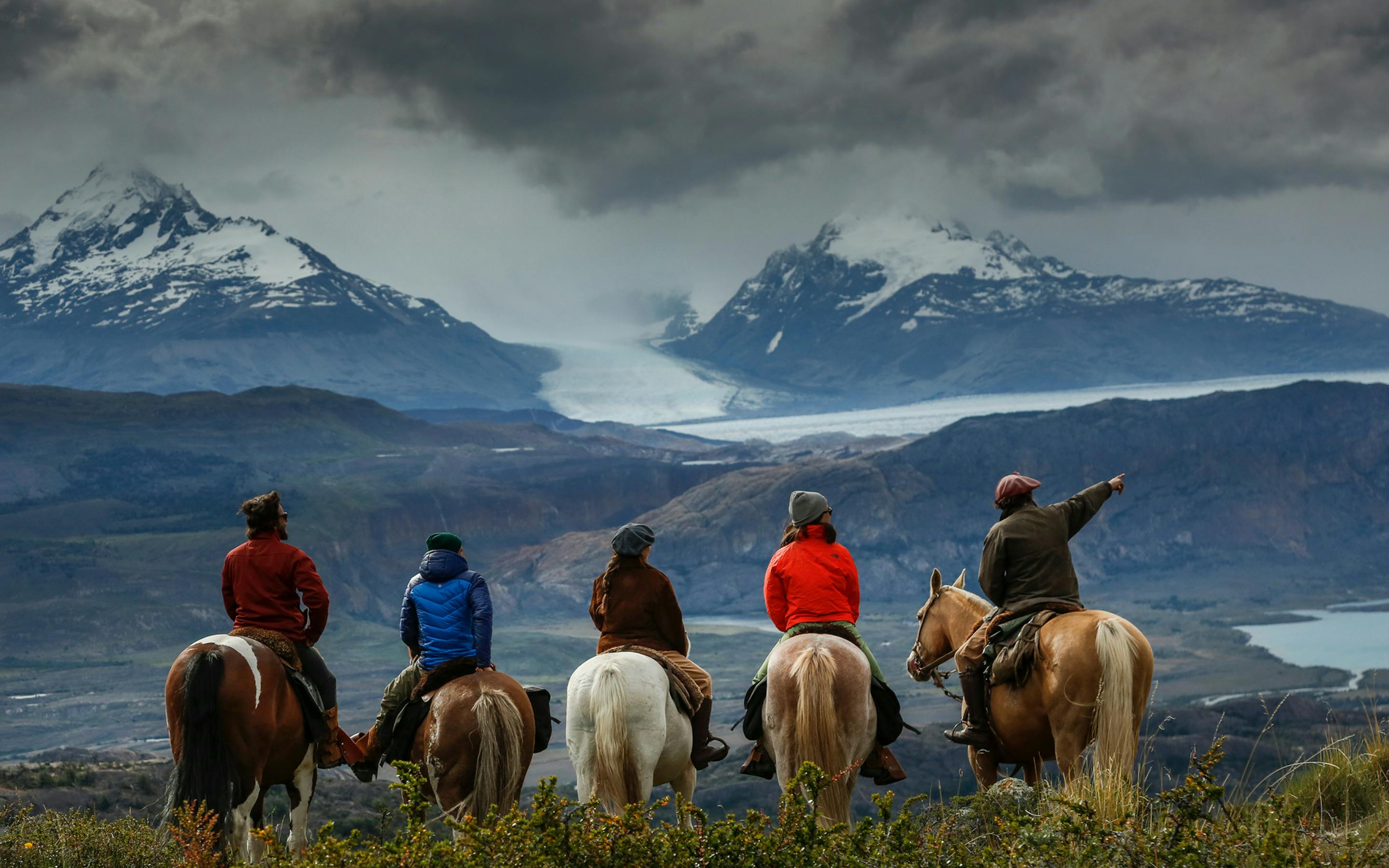 Riders on horseback cross open plains toward snow-dusted mountains beneath heavy clouds and misty ridgelines.