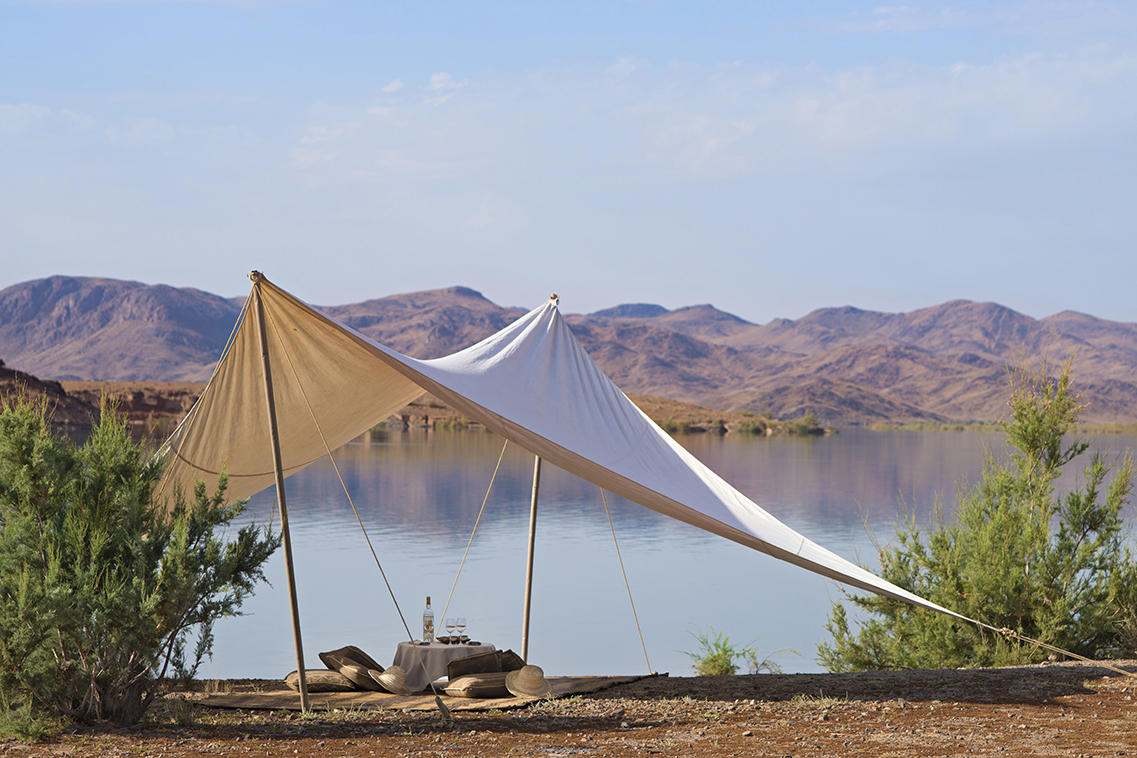 A shaded canvas tent sits beside a calm lagoon, with rocky desert hills and clear blue sky in the distance.