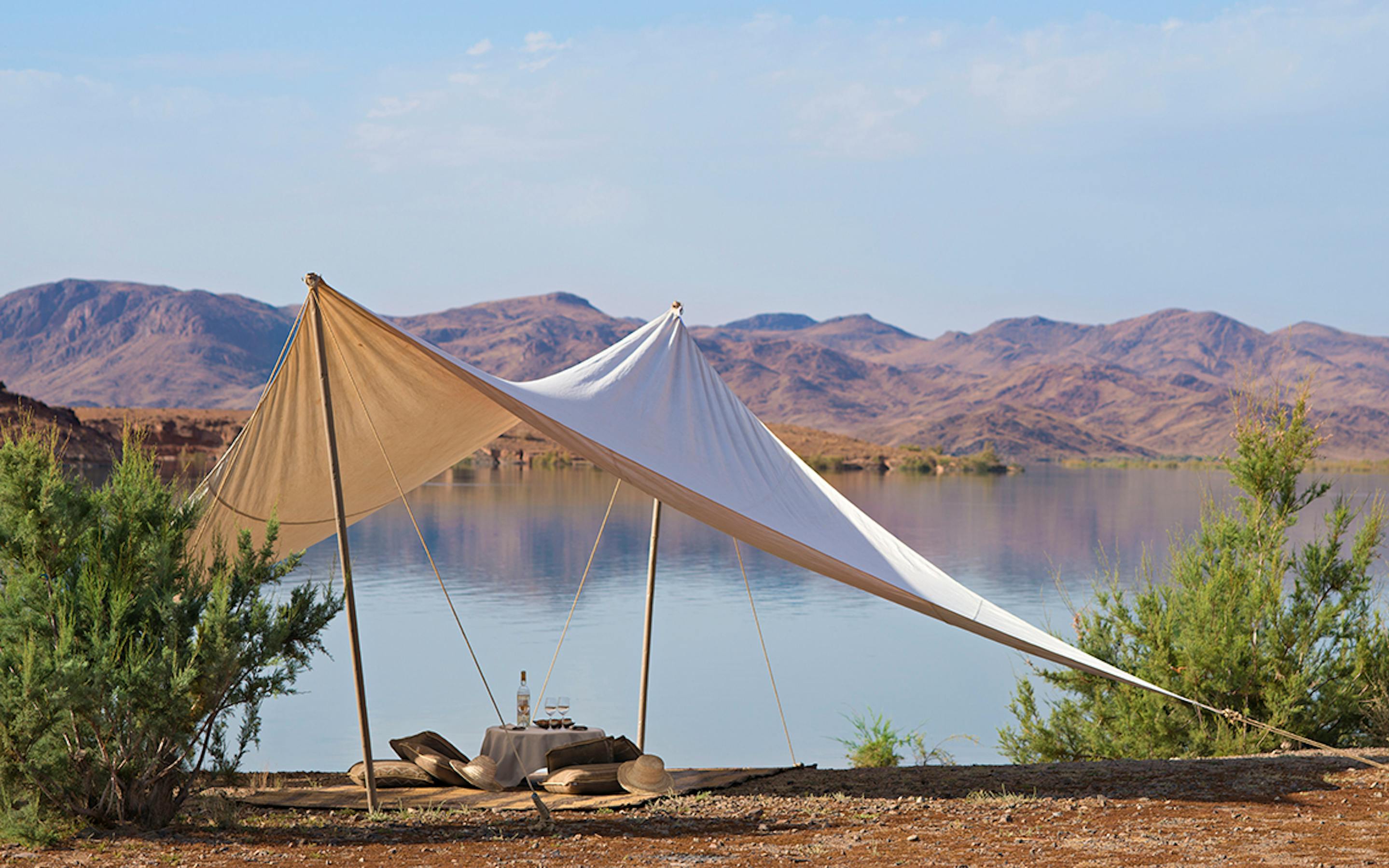 A shaded canvas tent sits beside a calm lagoon, with rocky desert hills and clear blue sky in the distance.