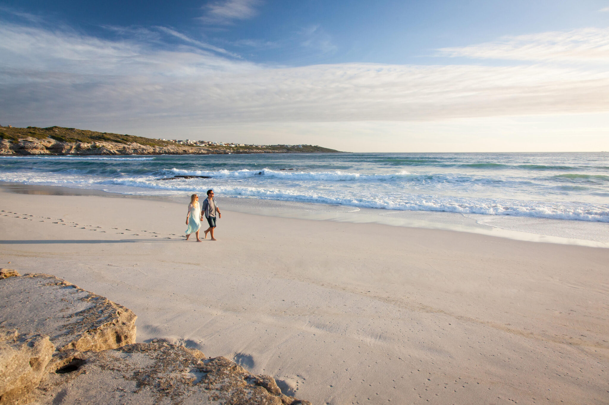 Two people walk along a wide sandy beach at the waterline as gentle waves roll in, with low cliffs in the distance.