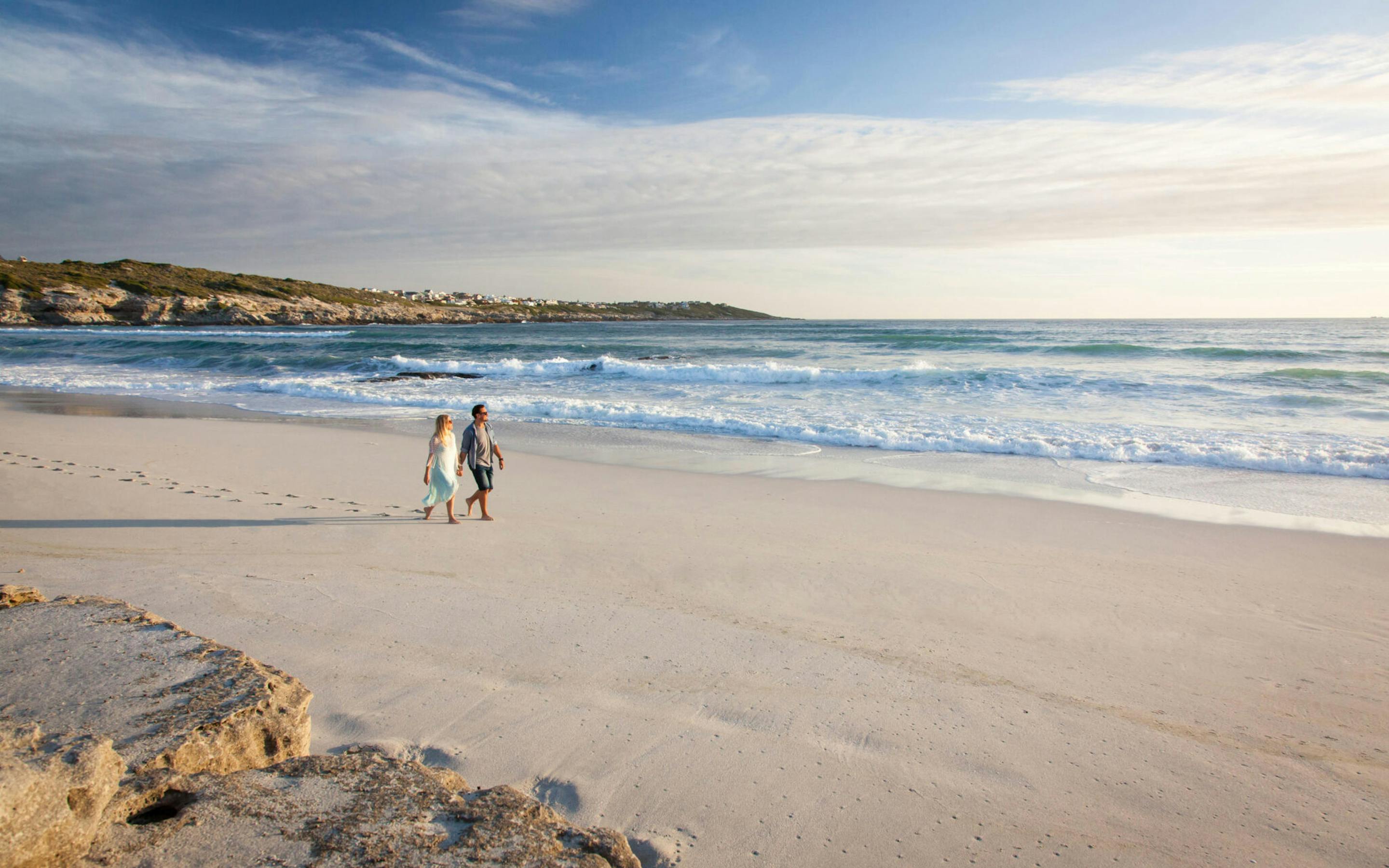 Two people walk along a wide sandy beach at the waterline as gentle waves roll in, with low cliffs in the distance.